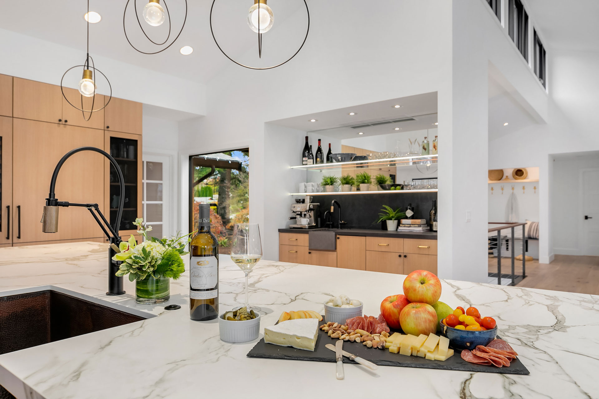 A modern kitchen with light wooden cabinets, a marble island, and black fixtures. The island features a wine bottle, a filled glass, a bouquet of flowers, and a serving tray with cheese, nuts, apples, and cherry tomatoes. Pendant lights hang above the island.