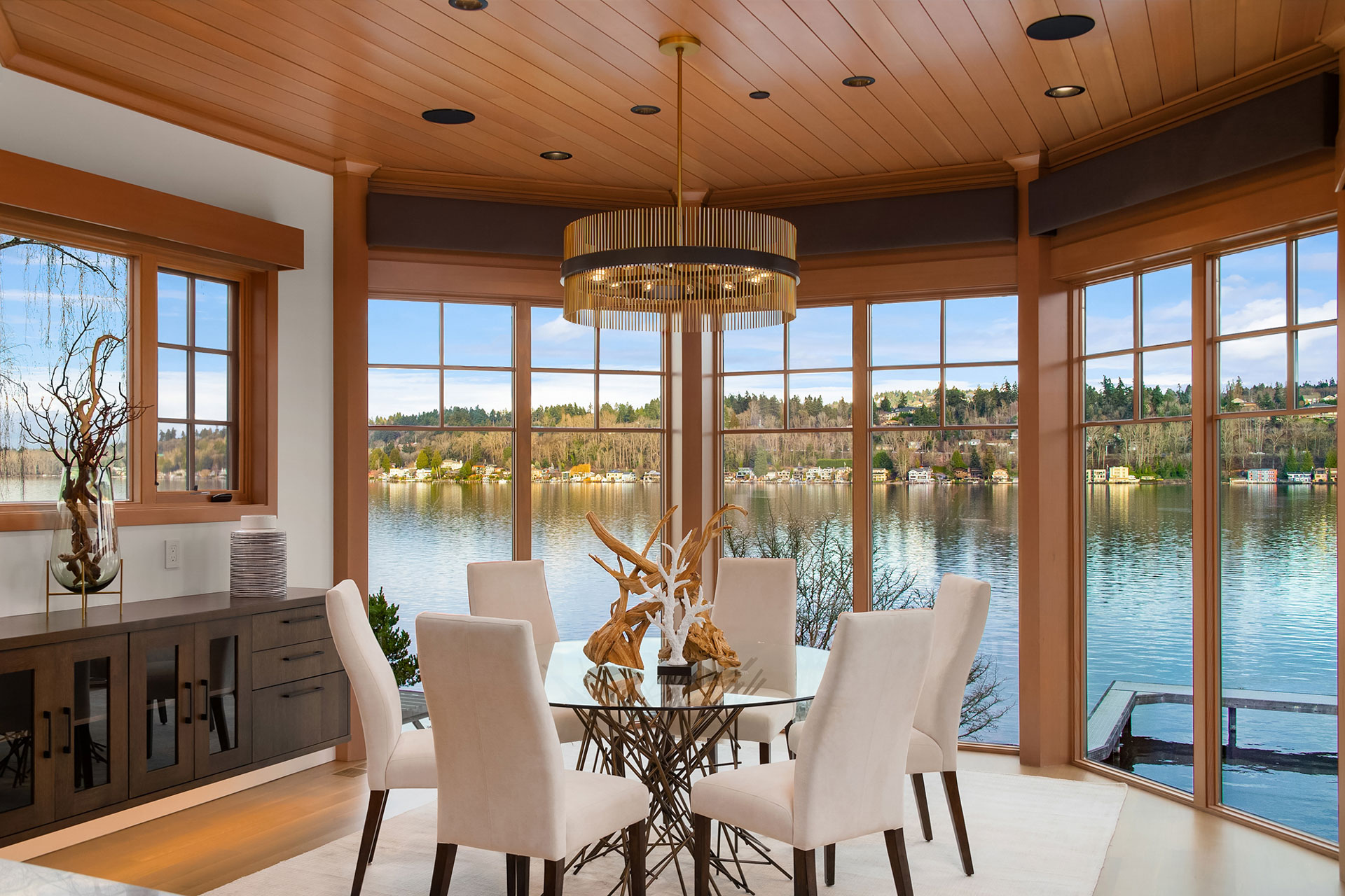 A dining room with large windows showcasing a scenic lake view. The room features a round glass table with a decorative branch centerpiece, surrounded by six white upholstered chairs. The ceiling is wooden with recessed lighting, and a modern chandelier hangs above the table.