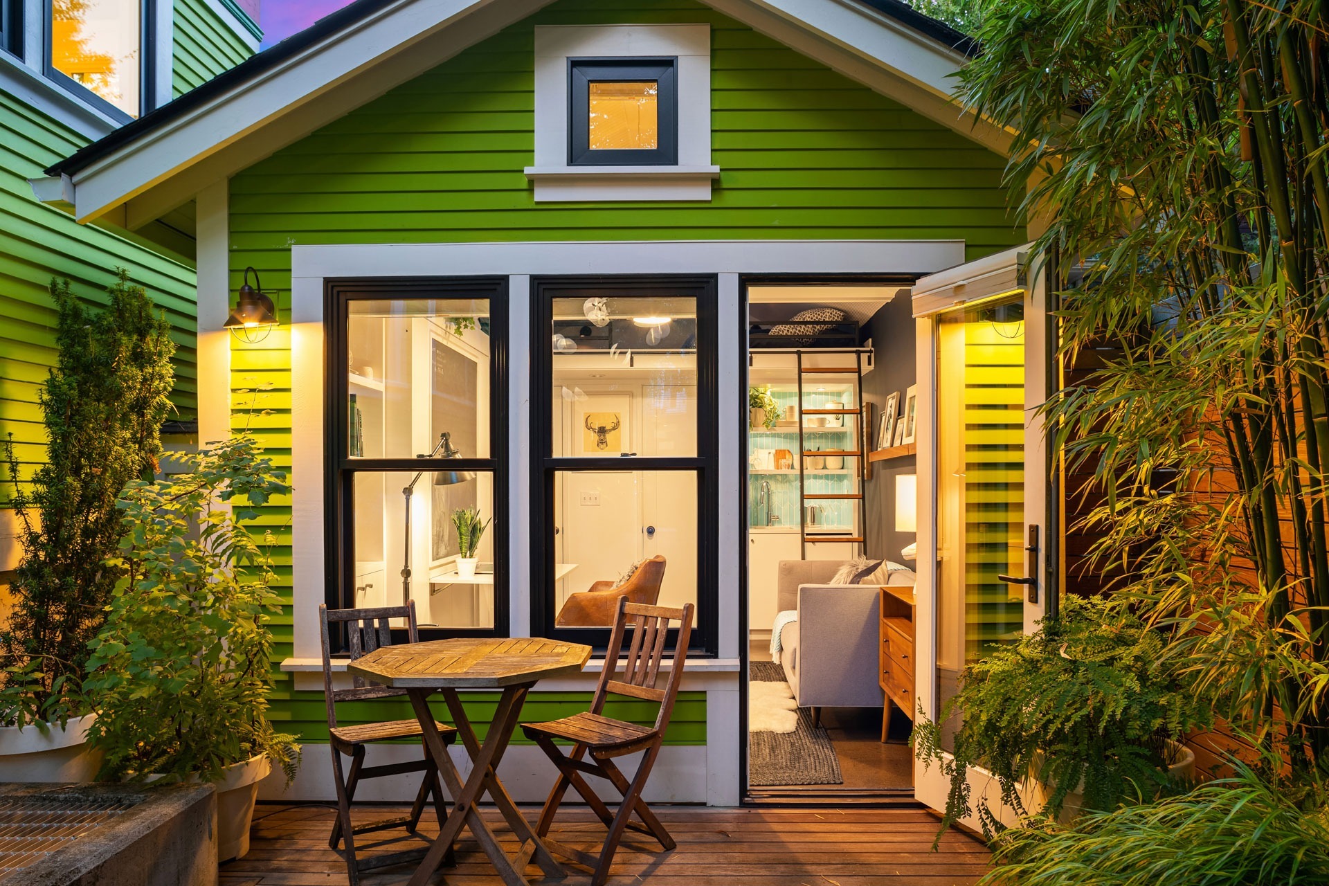 A cozy green house with white trim, warmly lit, featuring a small patio area with a round wooden table and two chairs. The patio is adorned with potted plants and tall bamboo, and through the open door, a stylishly furnished living room is visible.