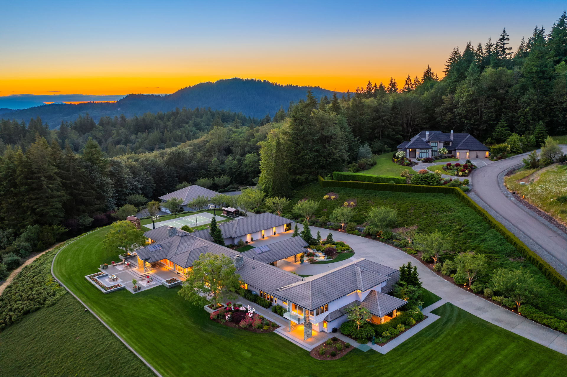 Aerial view of two luxury homes surrounded by lush greenery at sunset. The main home features extensive landscaping, a circular driveway, and ample outdoor lighting. A secondary home is located on an elevated area nearby. Rolling hills and forests complete the scenic backdrop.