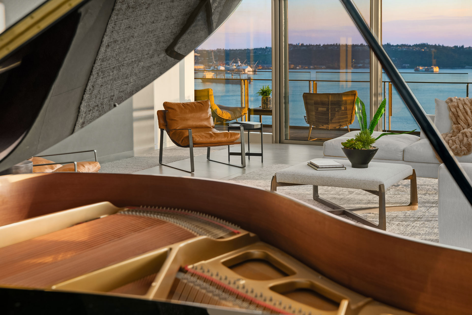 A grand piano in the foreground with an open lid, set in a modern living room featuring leather chairs, a coffee table with a plant, and a large window offering a view of a serene waterfront scene with boats and distant hills.