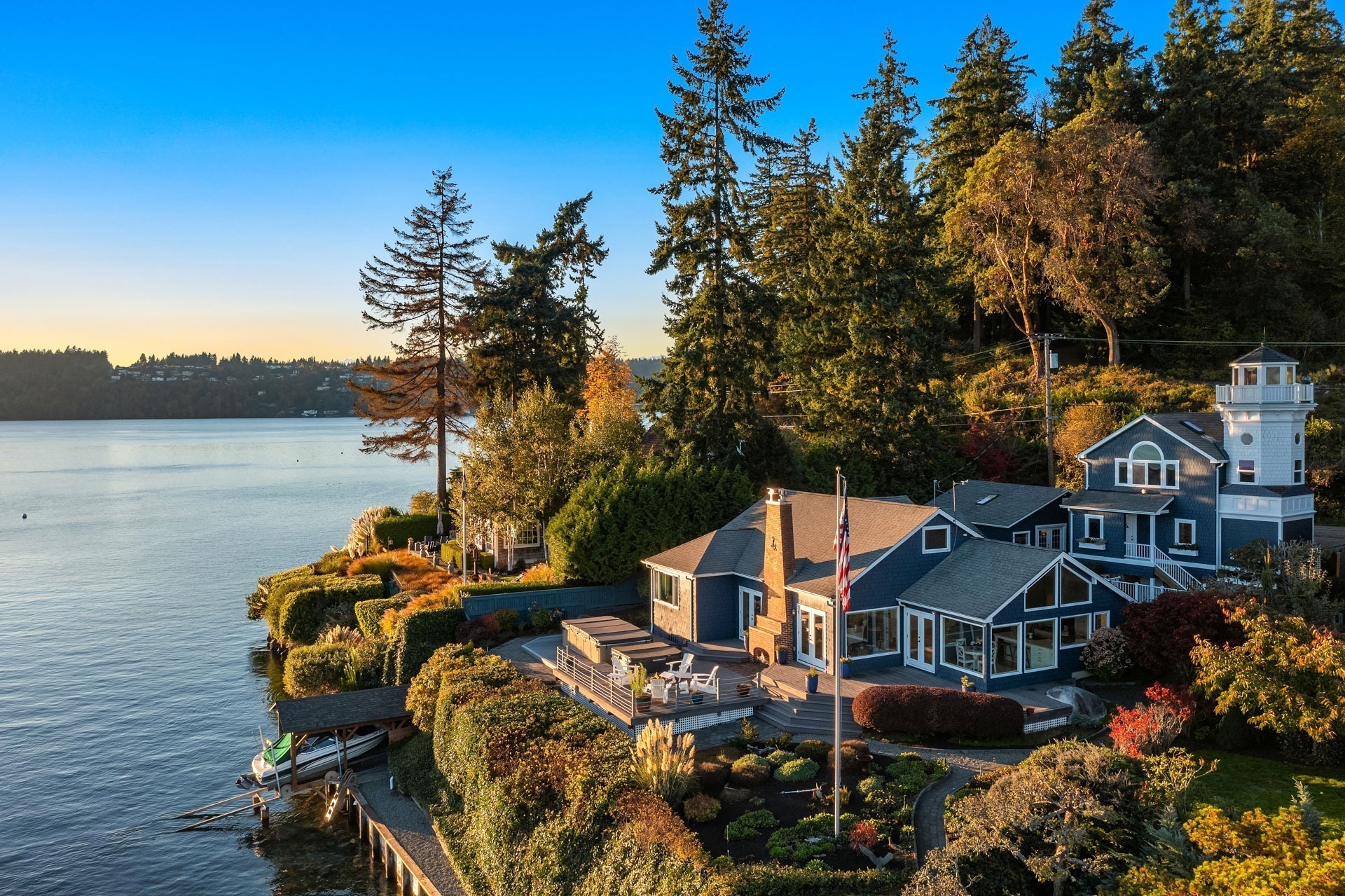 Aerial view of a lakeside property with a large house surrounded by lush greenery and colorful landscaping. The house has multiple stories, several large windows, and a flagpole visible in the yard. A small dock extends into the calm lake, reflecting the clear blue sky.