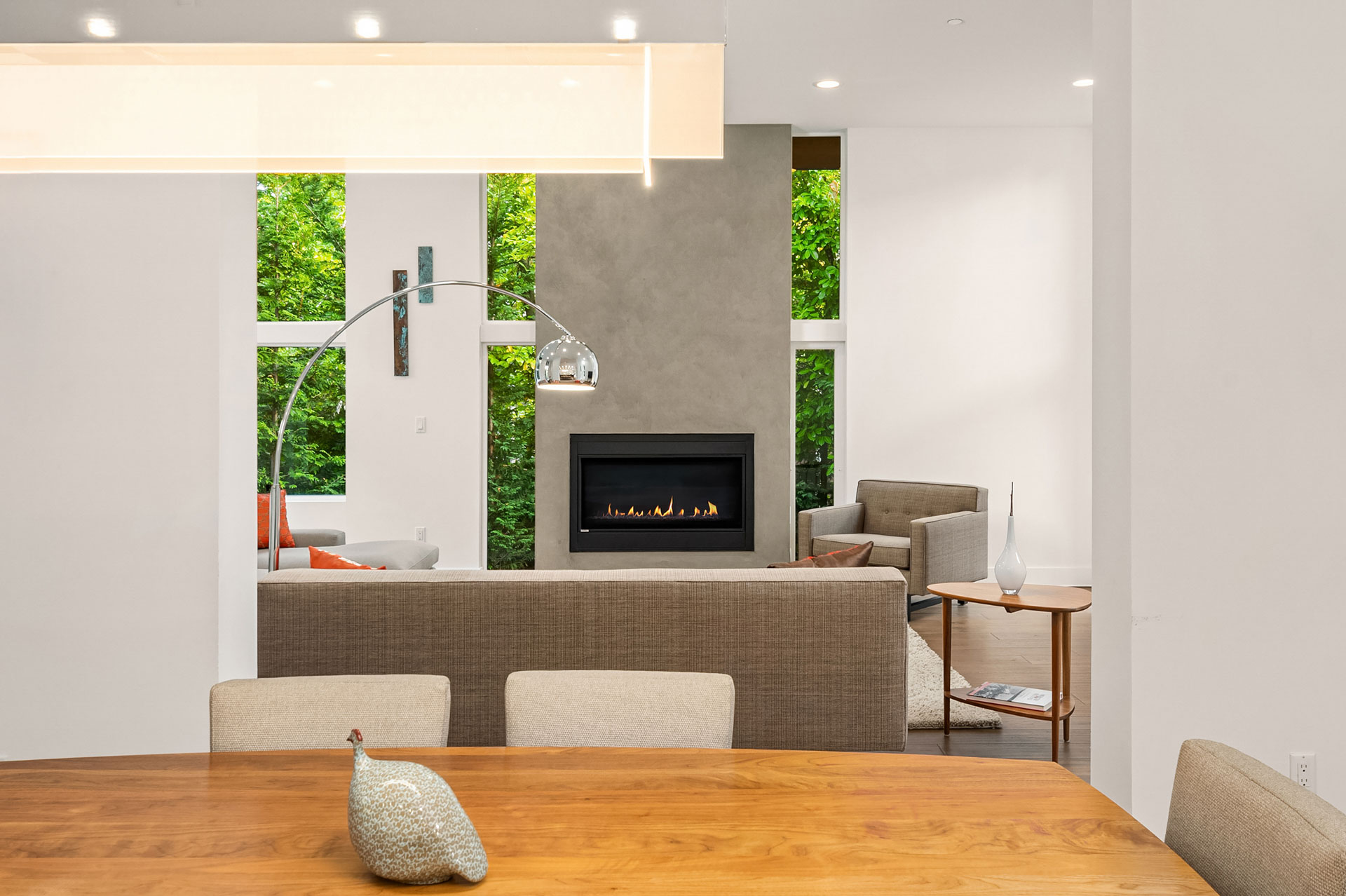 A modern living room with a minimalist design features a gray sectional sofa, an arc floor lamp, a wall-mounted fireplace, and large windows showcasing lush green trees. A wooden table with beige chairs is in the foreground.