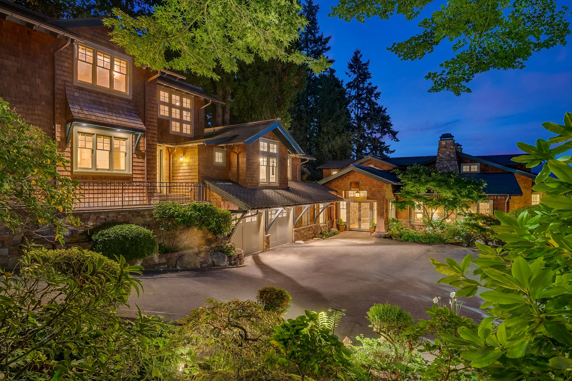 A spacious, rustic house at dusk surrounded by lush greenery. The house features large, illuminated windows, wood siding, and a multi-gabled roof. The driveway leads to a well-lit entrance, with a forested area and evening sky in the background.