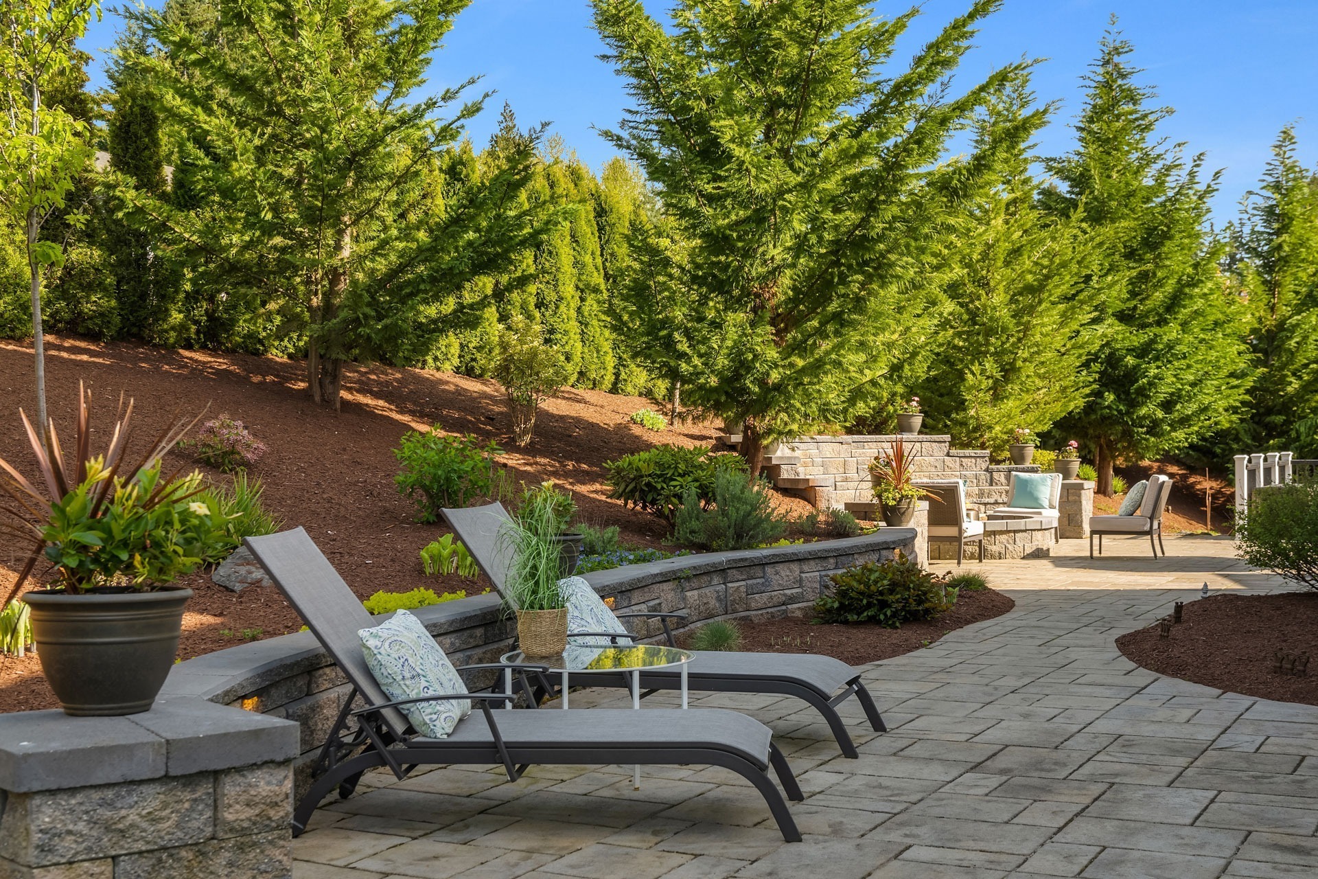 A landscaped backyard with two gray lounge chairs on a paved patio, surrounded by lush evergreen trees. A stone retaining wall and potted plants add to the decor. In the background, there is a seating area with outdoor furniture under the blue sky.