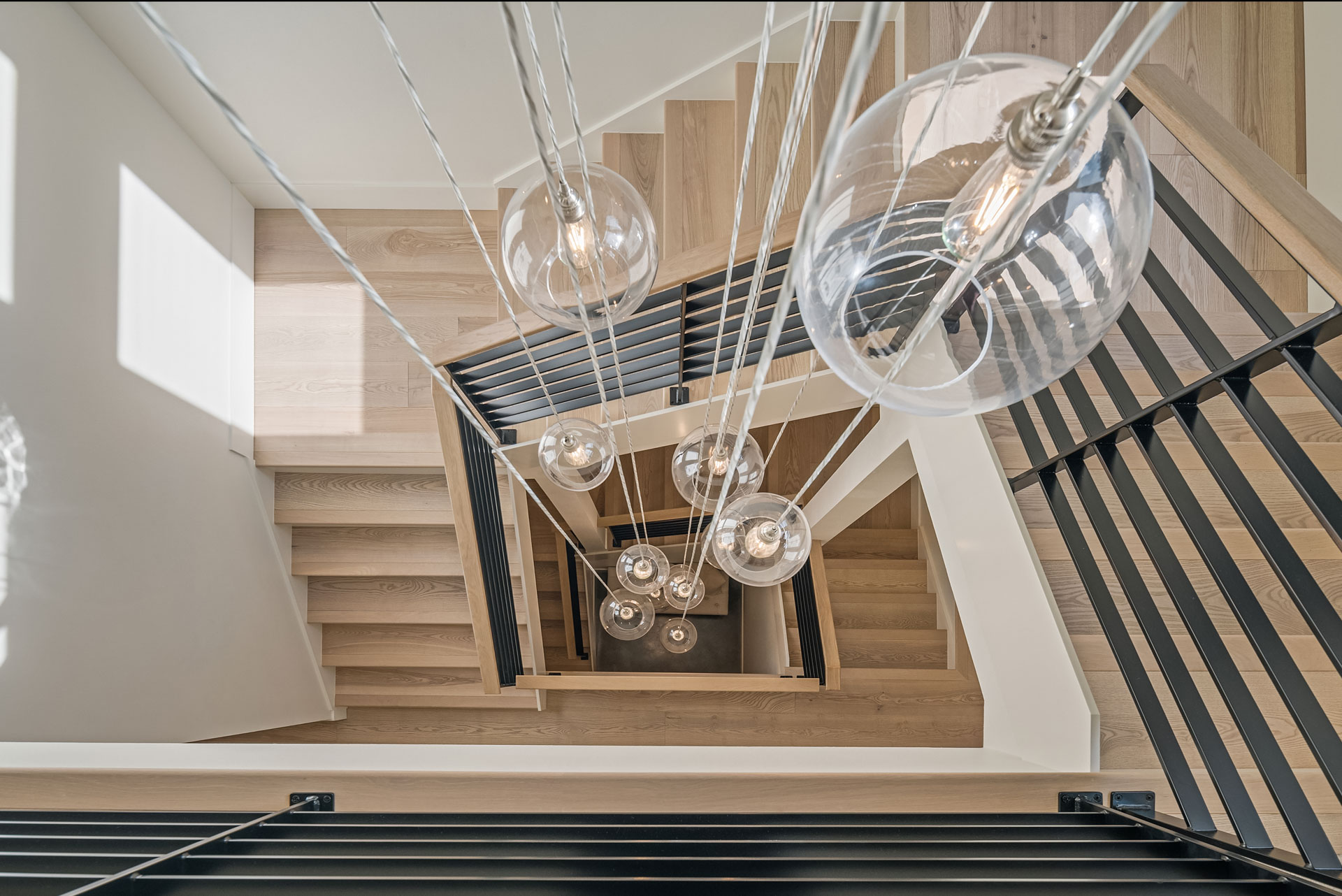 A top-down view of a modern spiral staircase with light wooden steps and black metal railings. Multiple hanging glass light fixtures with exposed bulbs are suspended in the center of the stairwell, creating a contemporary and visually striking design.