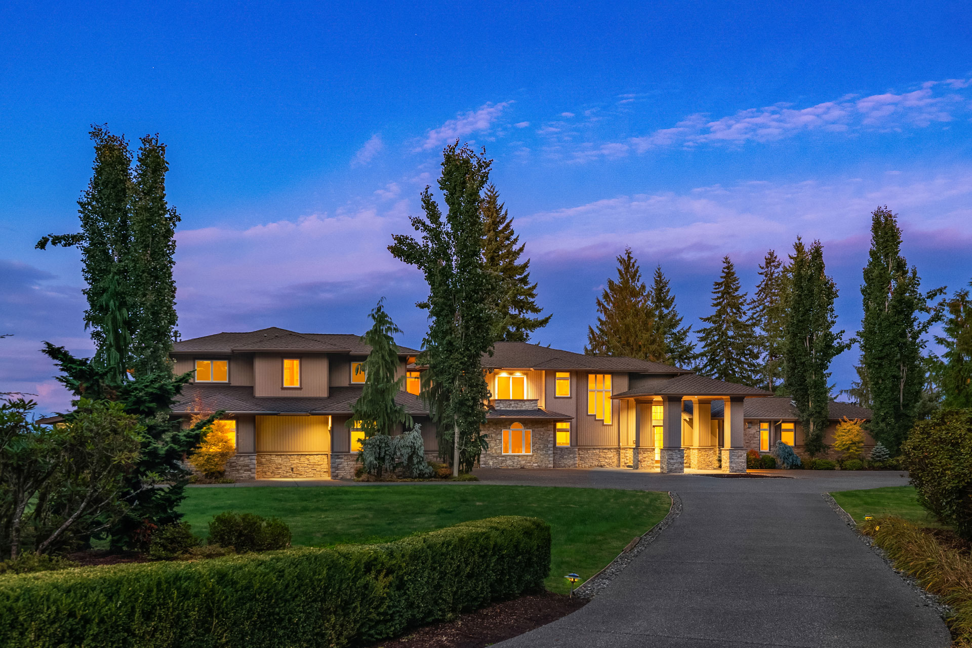 A large, elegant two-story house with warm interior lighting is surrounded by landscaped greenery, tall trees, and a carefully manicured lawn. A wide driveway leads up to the entrance, and the sky is clear with a beautiful gradient of colors at twilight.