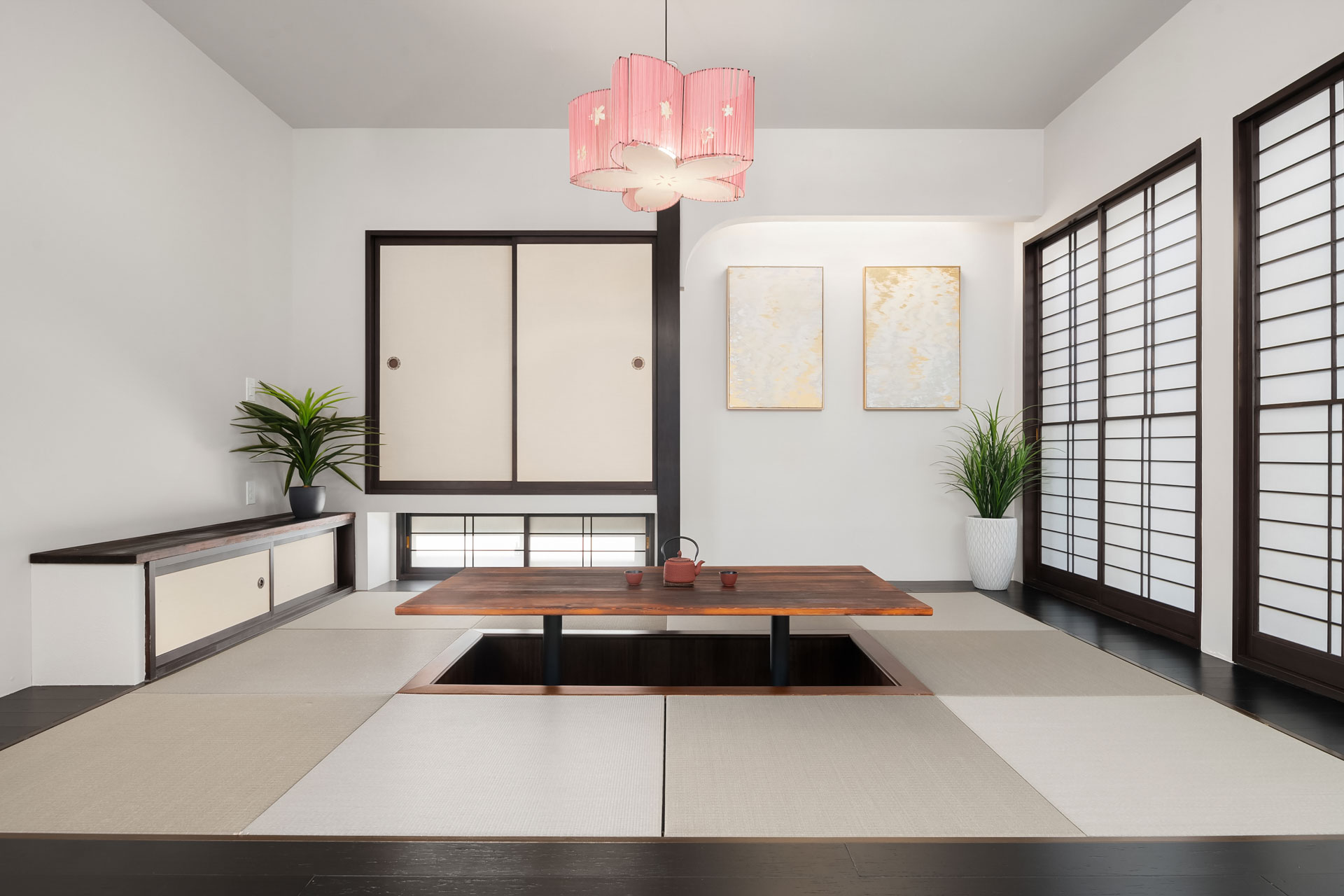 A minimalistic Japanese-style room with a sunken wooden table in the center, surrounded by tatami mats. Shōji sliding doors are on the right, and a white wall with two art pieces. A potted plant is to the left beneath the pink ceiling light.