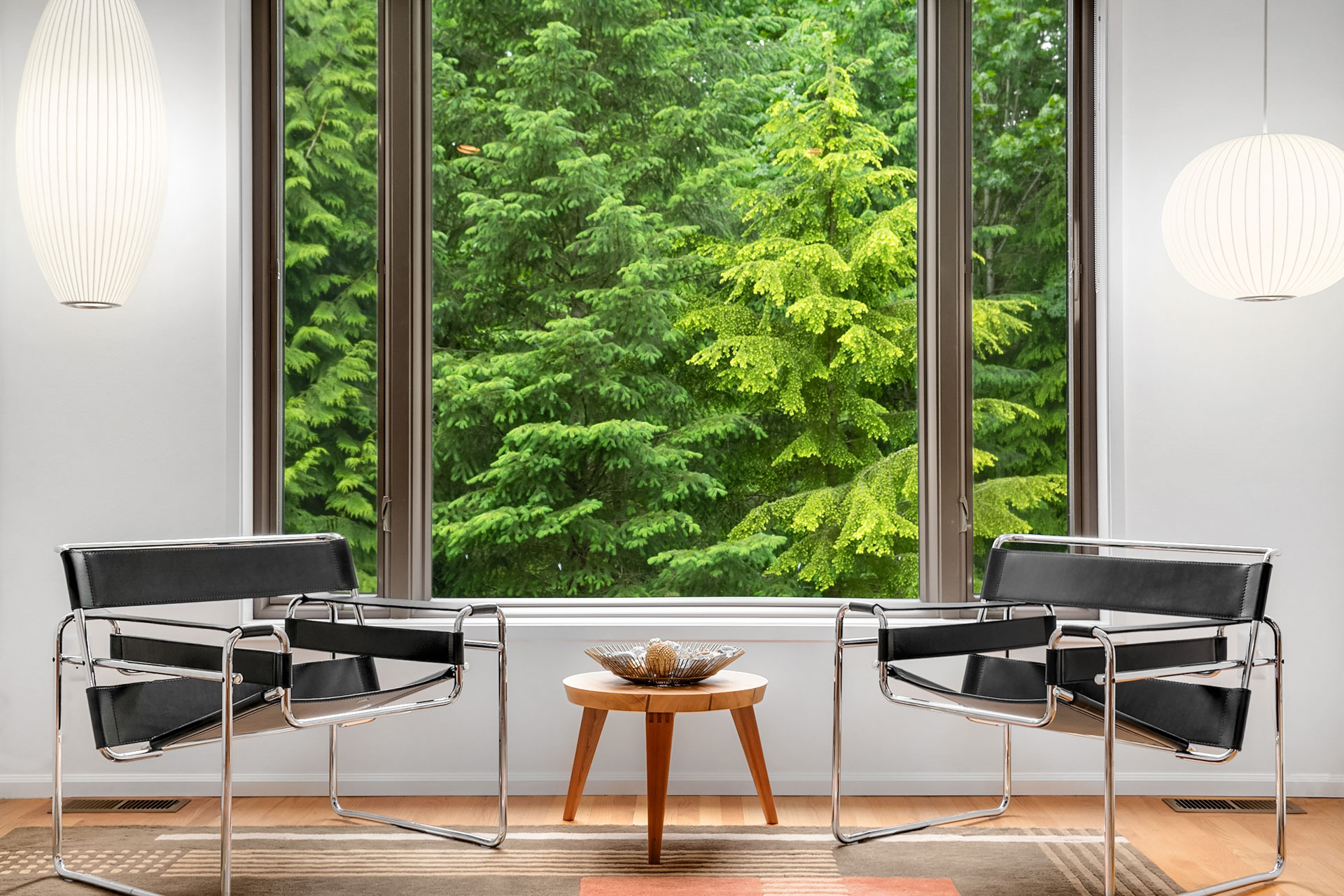 A modern, minimalist room with two black and chrome chairs facing each other, separated by a small wooden table with a decorative bowl. Large windows reveal lush green trees outside. Two white pendant lights hang from the ceiling, casting a soft glow.