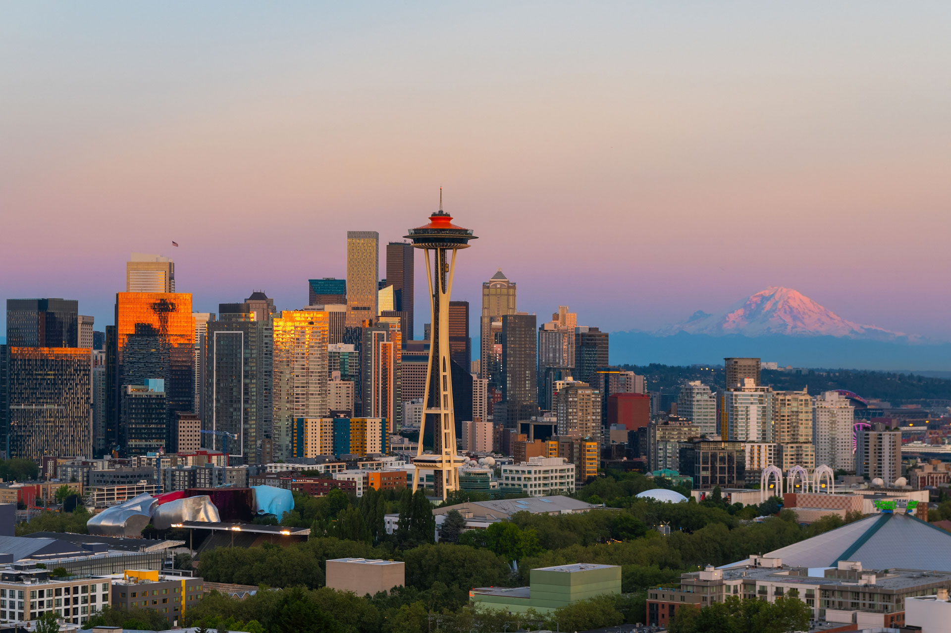 A scenic view of the Seattle skyline during sunset with the iconic Space Needle in the foreground. The city's high-rise buildings reflect a golden hue, and to the right, Mount Rainier stands majestically, partially shrouded in soft pink and purple tones.