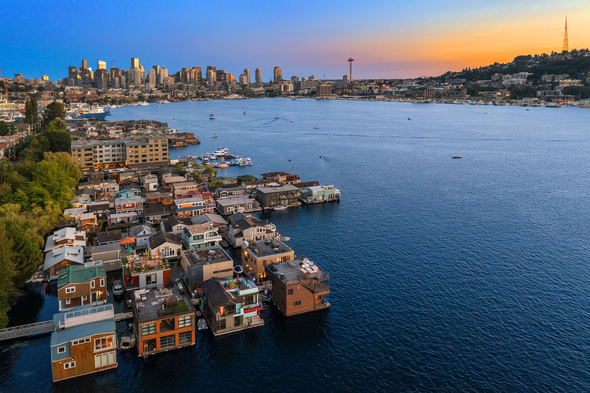 Aerial view of Seattle during sunset, with houseboats in the foreground on Lake Union and the downtown skyline in the distance. The Space Needle is visible on the right, and boats dot the water. The sky transitions from deep blue to warm orange hues.