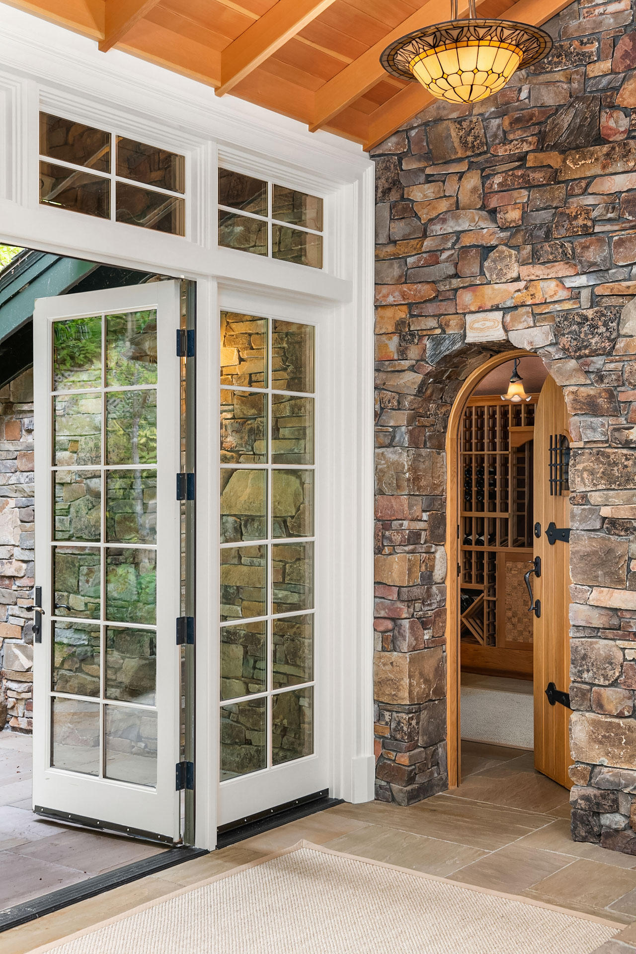 A rustic entryway features a stone wall with an arched wooden door leading to a wine cellar. Adjacent is a large glass-paned double door with white framing, opening to an outdoor space. The ceiling is wooden, and an elegant light fixture hangs above.