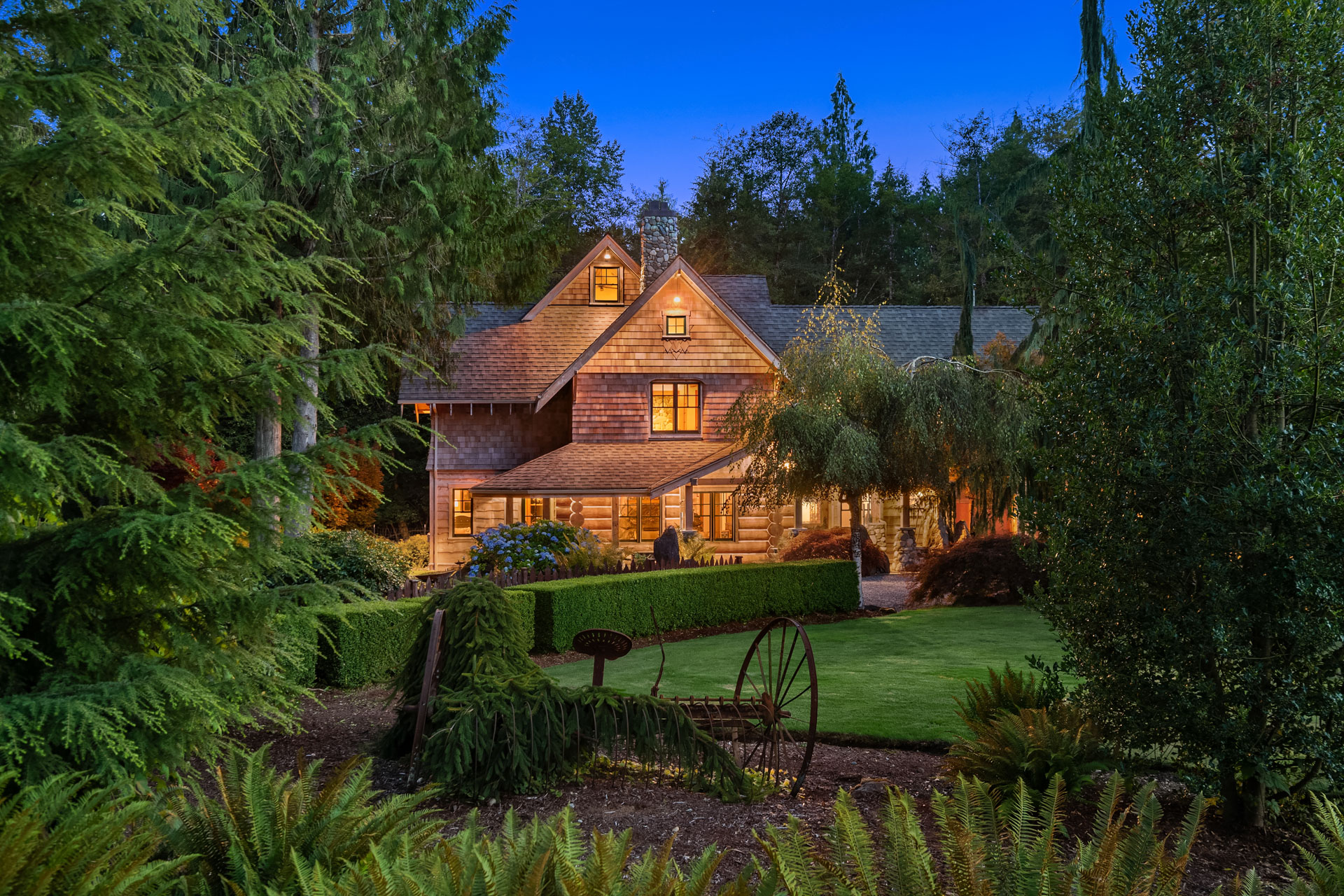 A charming, rustic two-story house nestled among dense greenery and tall trees. The house has warm, glowing lights emanating from its windows, a wooden exterior, and a neatly landscaped garden with a vintage wagon as a decorative feature. The sky is a deep twilight blue.