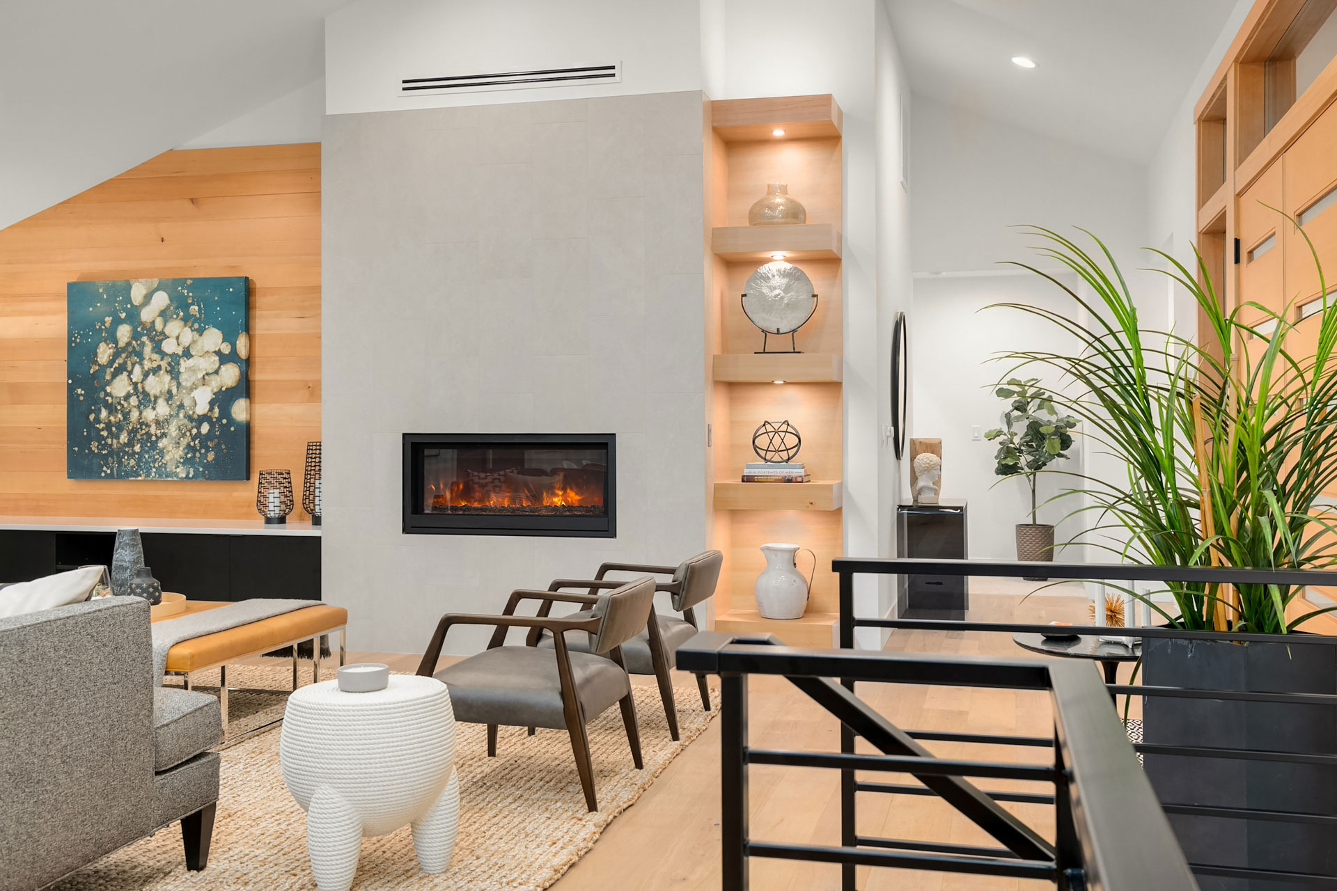 A modern living room with a fireplace, sleek seating, and light wooden shelving displaying decor items. A large, vibrant painting hangs on the wooden wall. A mix of contemporary furniture and lush greenery add to the bright and open ambiance.