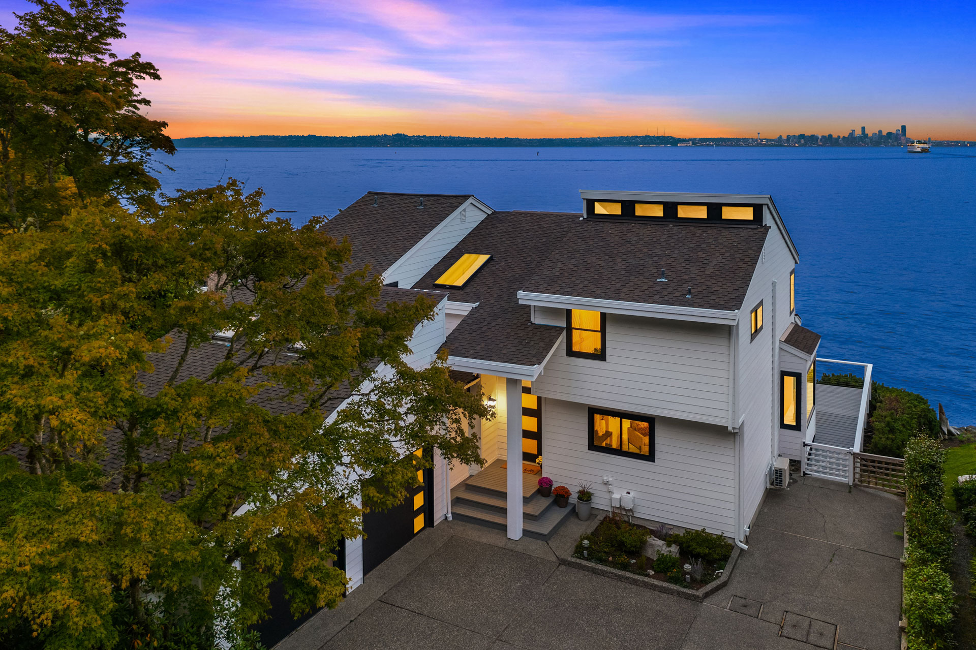 A modern, white multi-story house with large windows sits by the water's edge, featuring a scenic view of a city skyline across the body of water at sunset. The house is surrounded by greenery and has a spacious driveway in front.