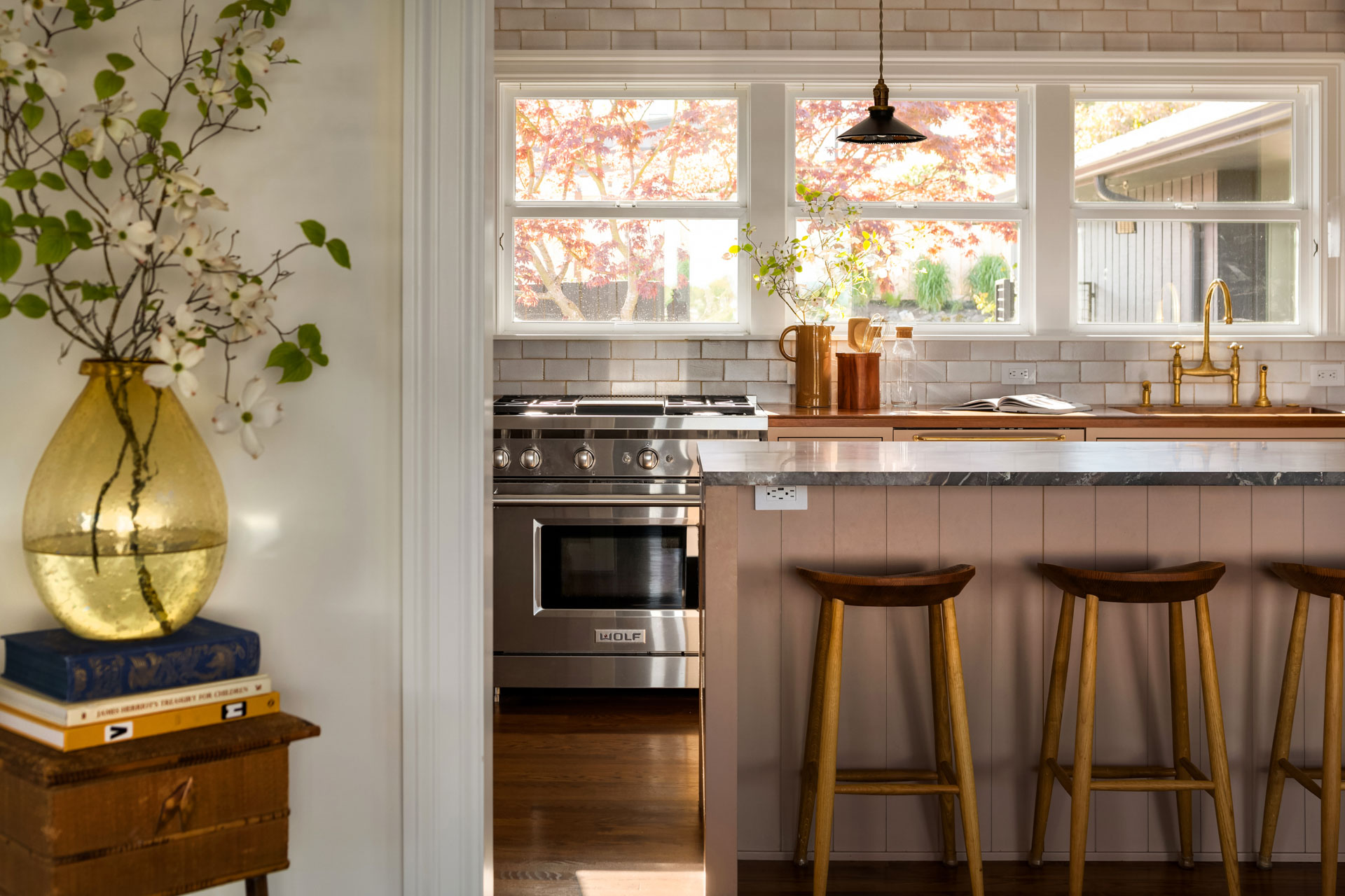 A cozy kitchen with a light beige and wood color scheme. There's a stainless steel stove, a marble-topped island with three wooden stools, a sink with brass fixtures, and a vase with flowers by the window. A potted plant and books sit on a wooden side table to the left.