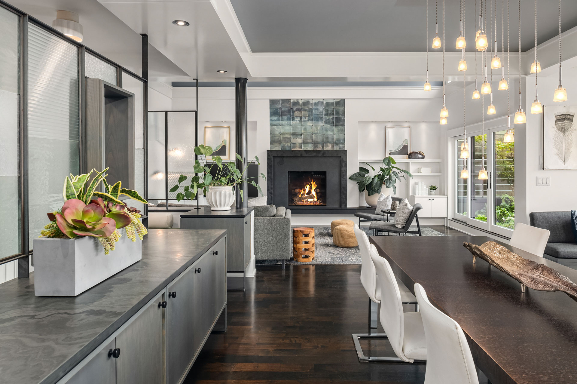 A modern, open-concept living and dining area with dark hardwood flooring. The room features white walls, a cozy fireplace, hanging pendant lights, and large windows. Potted plants and contemporary furniture, including white chairs and a dark dining table, add to the aesthetic.