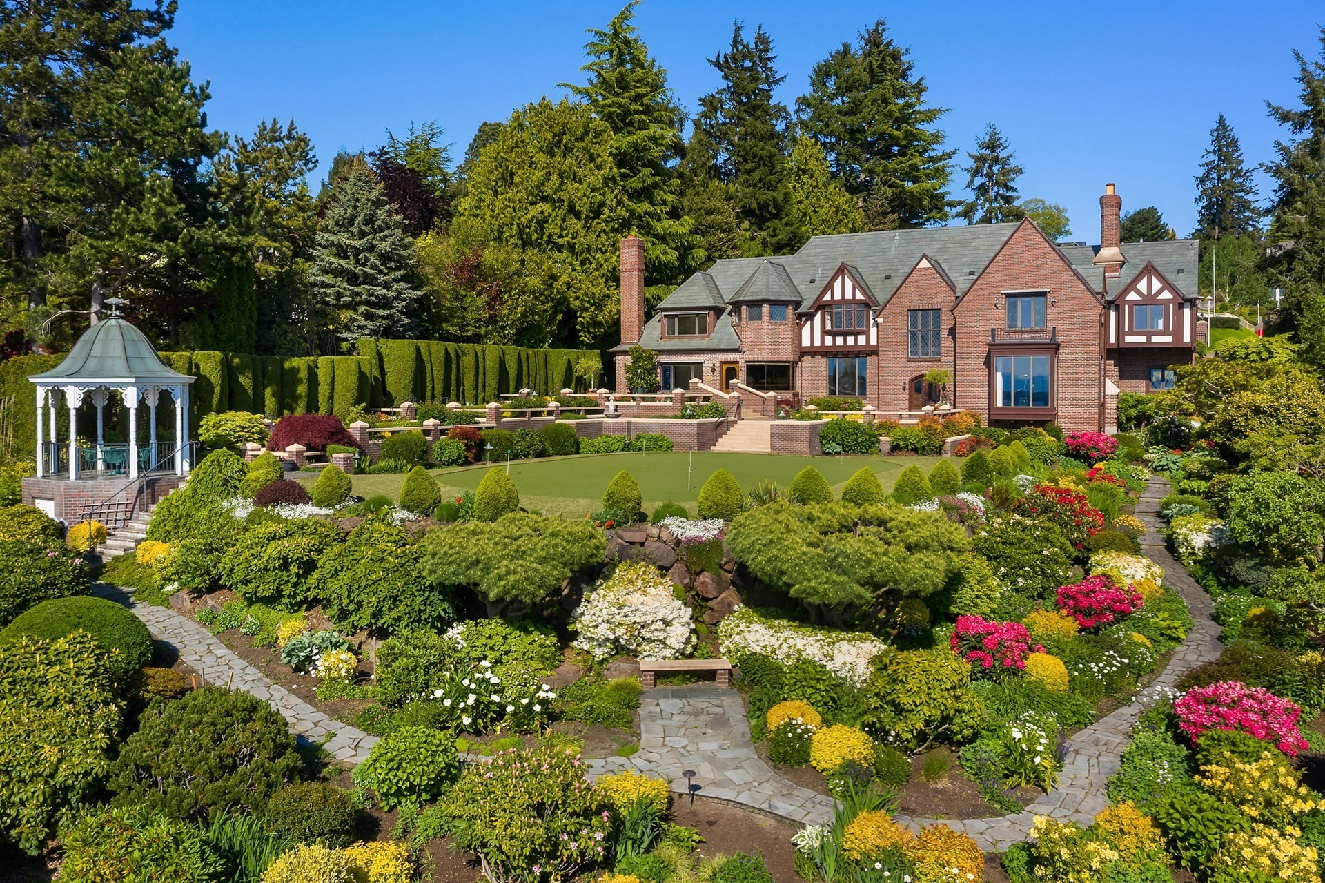 A large, luxurious brick house with multiple chimneys and gabled roofs stands in the background. The foreground features a lush, landscaped garden with colorful flowers, neatly trimmed hedges, and a stone pathway. On the left side, there is a white gazebo.