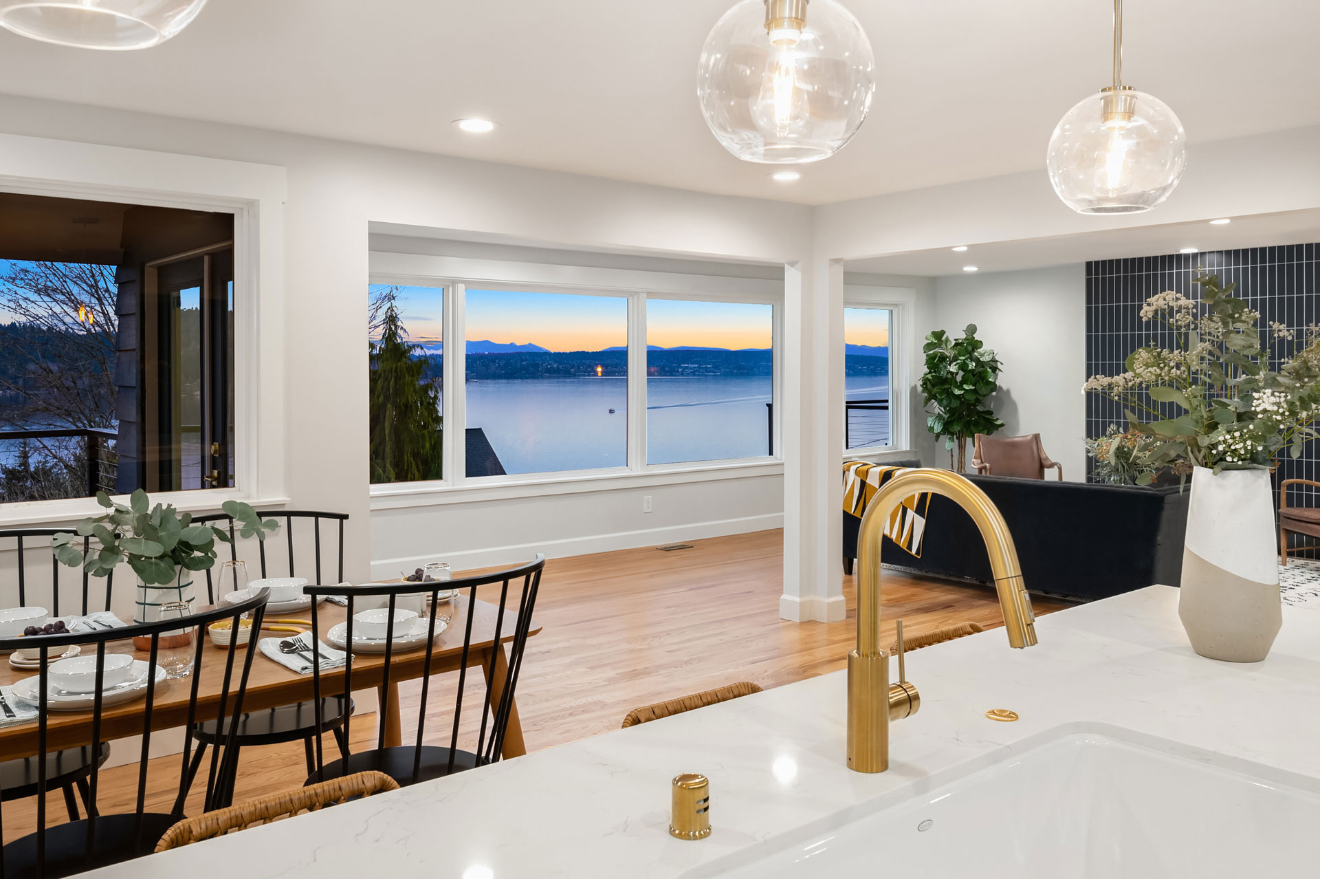 A modern, open-concept dining and living space with large windows offering a sunset view over a lake. A wooden dining table is set for four, and the kitchen island features a gold faucet. The room is decorated with plants and contemporary light fixtures.