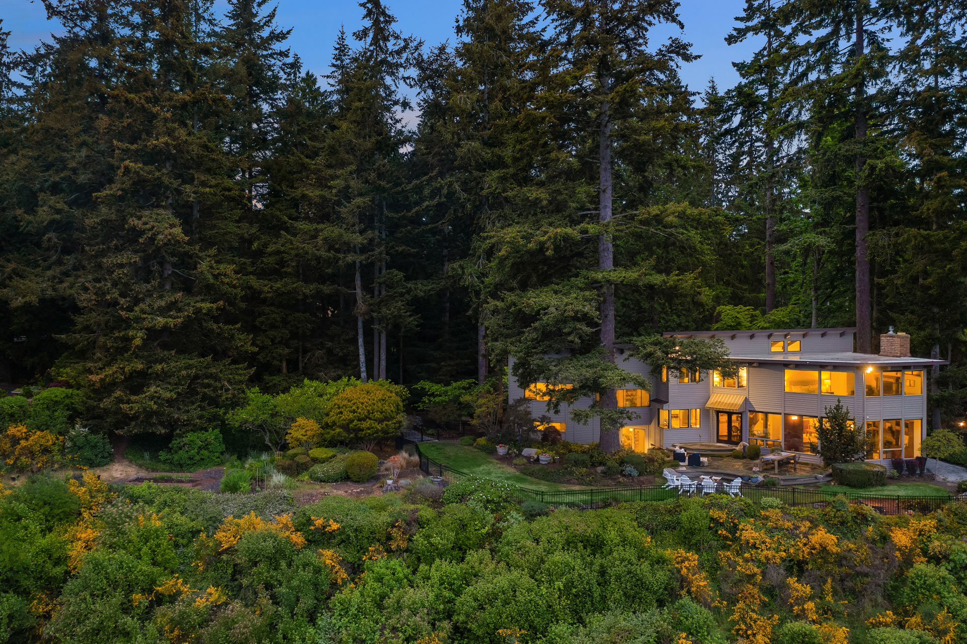 A large, two-story house with warm indoor lighting is nestled amidst tall trees and lush greenery at dusk. The property features a spacious outdoor patio with seating, surrounded by dense forest and mature landscaped gardens.