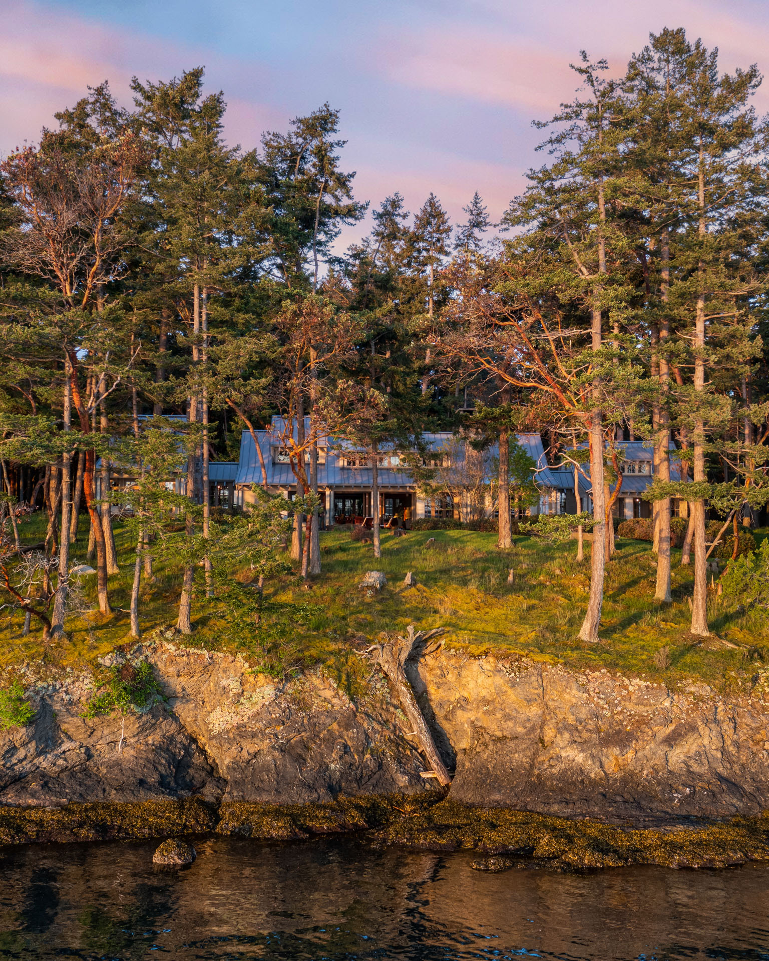 A house nestled among tall trees on a cliffside with rocky edges, overlooking calm waters. The setting sun casts a golden hue on the scene, highlighting the greenery and the tranquil atmosphere of the secluded home.