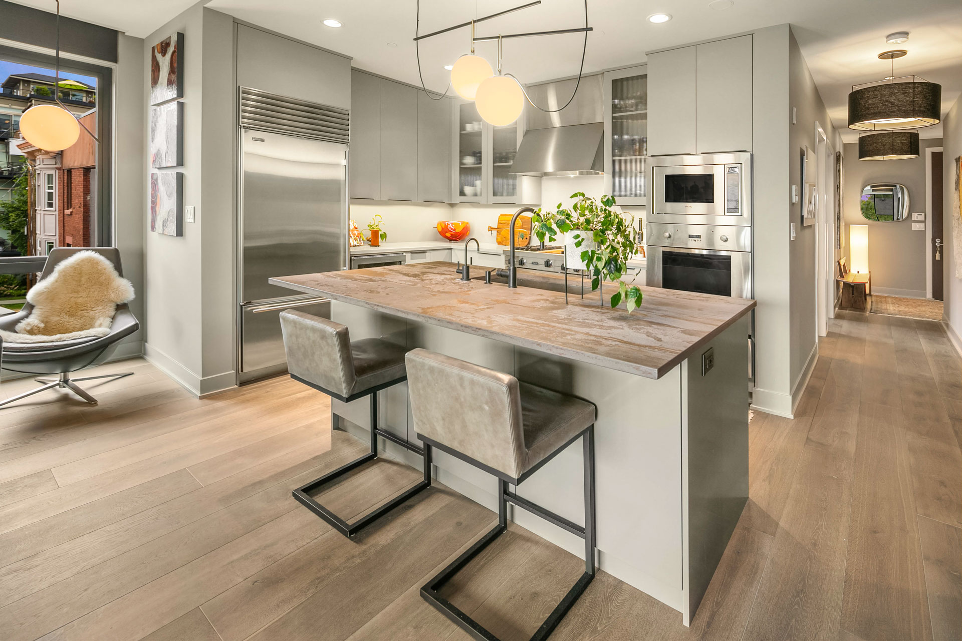 Modern kitchen with gray cabinetry, a large island with seating for two, stainless steel appliances, and wood flooring. Bright pendant lights hang above the island, and a cozy chair with a fur throw is positioned near a window with a view of a brick building.