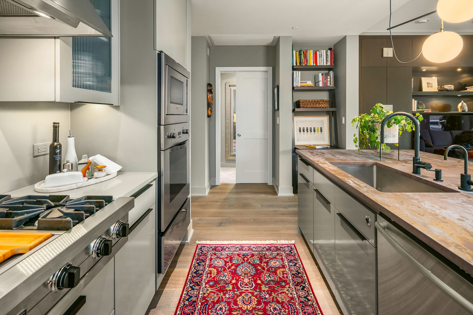 A modern kitchen with gray cabinets, stainless steel appliances, and a wooden countertop. A colorful red rug lies on the floor. There's a sink in the foreground, a stove and oven to the left, and a doorway leading to another room in the background.