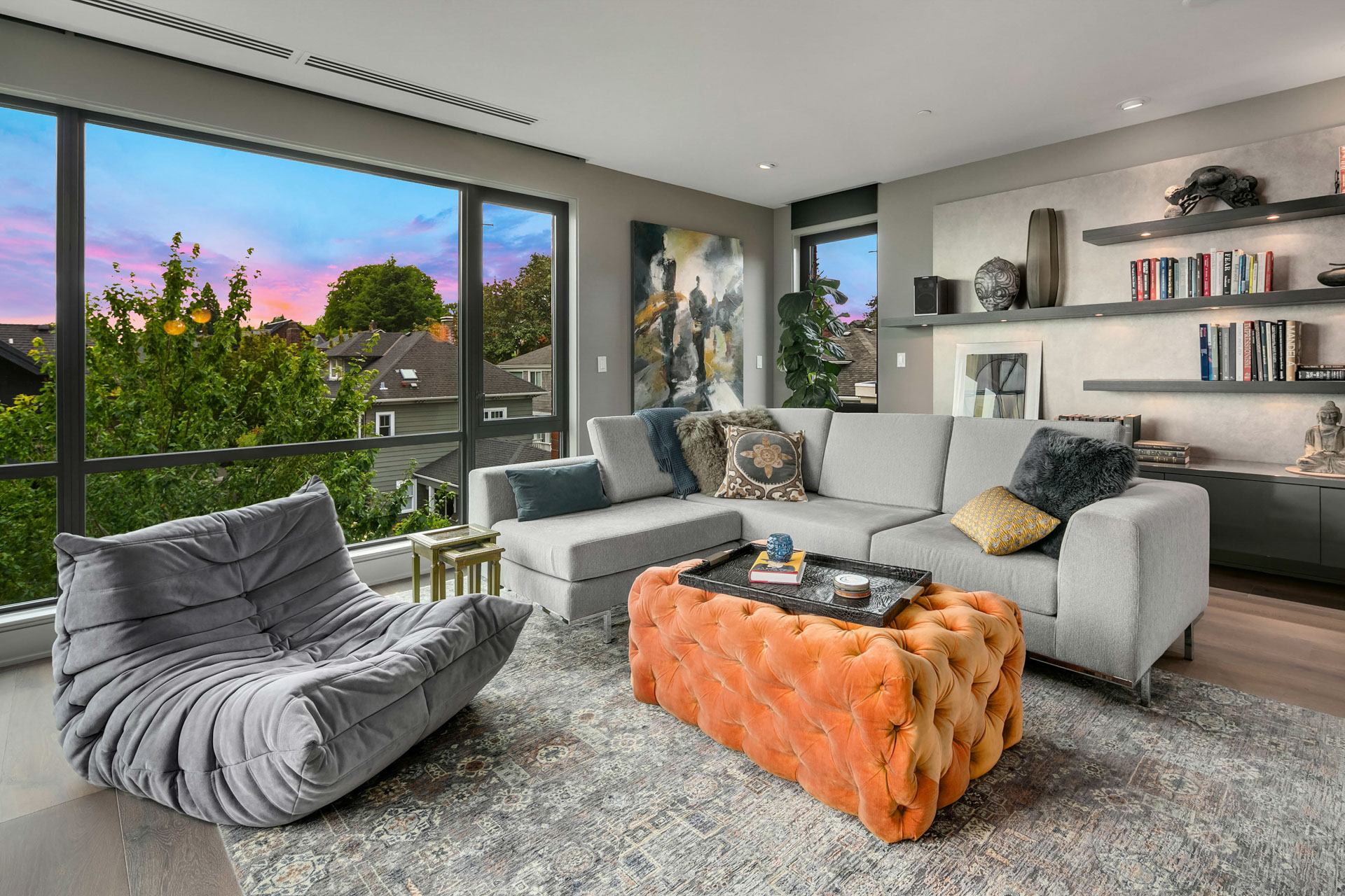 A modern living room featuring a large gray sectional sofa adorned with decorative pillows, a tufted orange ottoman used as a coffee table, and a cozy gray lounge chair. Large windows reveal a colorful sunset over a suburban neighborhood. Shelves with books and decor occupy the wall.