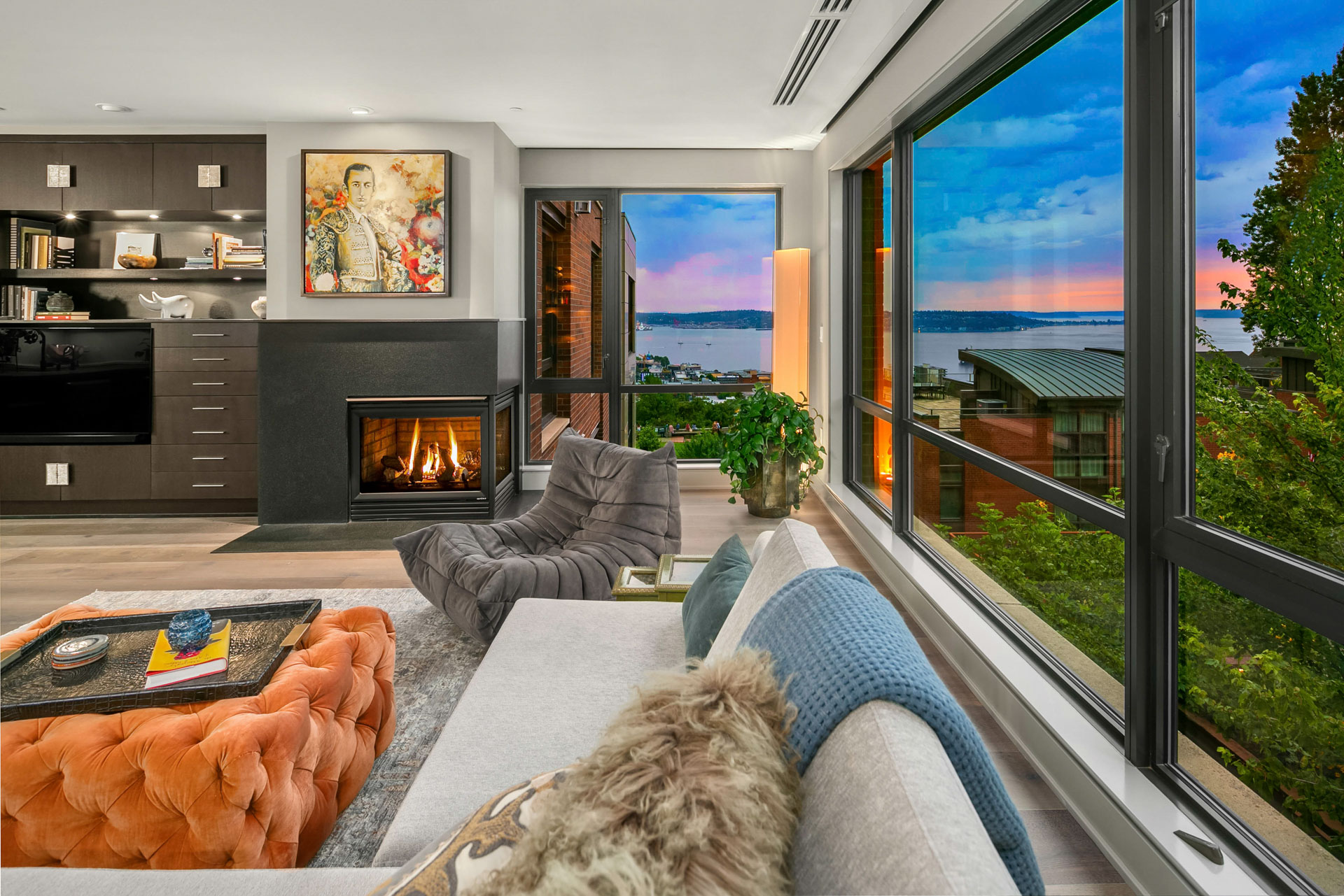 A modern living room featuring a large window with a sunset view over a body of water. The space includes a gray sofa, an orange tufted ottoman, a fireplace with a painting above it, and built-in shelves. A plush gray chair is near the window.