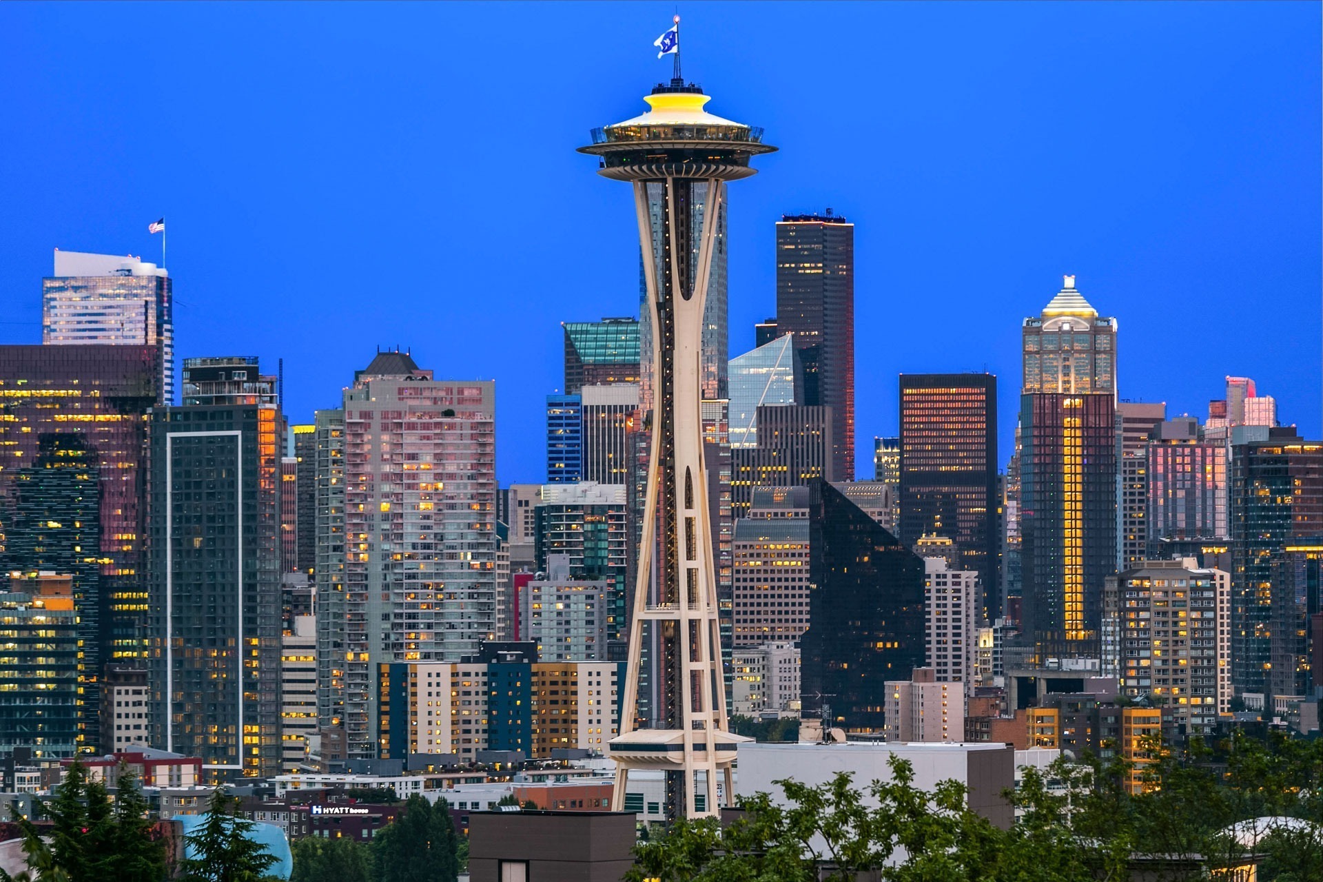 Seattle skyline at dusk featuring the Space Needle prominently in the center. The skyline is filled with various high-rise buildings illuminated against a deep blue sky, showcasing a mix of modern and classic architecture.