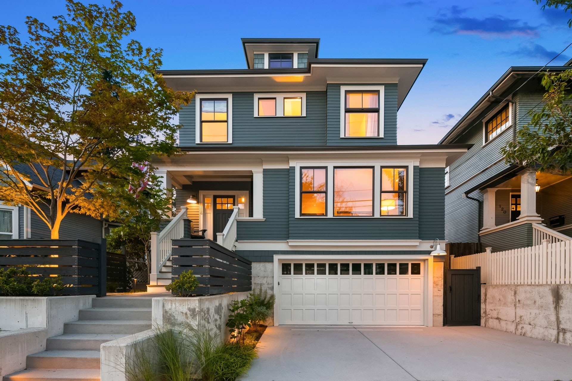 A two-story house with a modern design featuring a dark green exterior, large windows, and a spacious white garage. The house is framed by a tree on the left and a fence on the right. A staircase leads up to the entrance, flanked by minimalist landscaping.