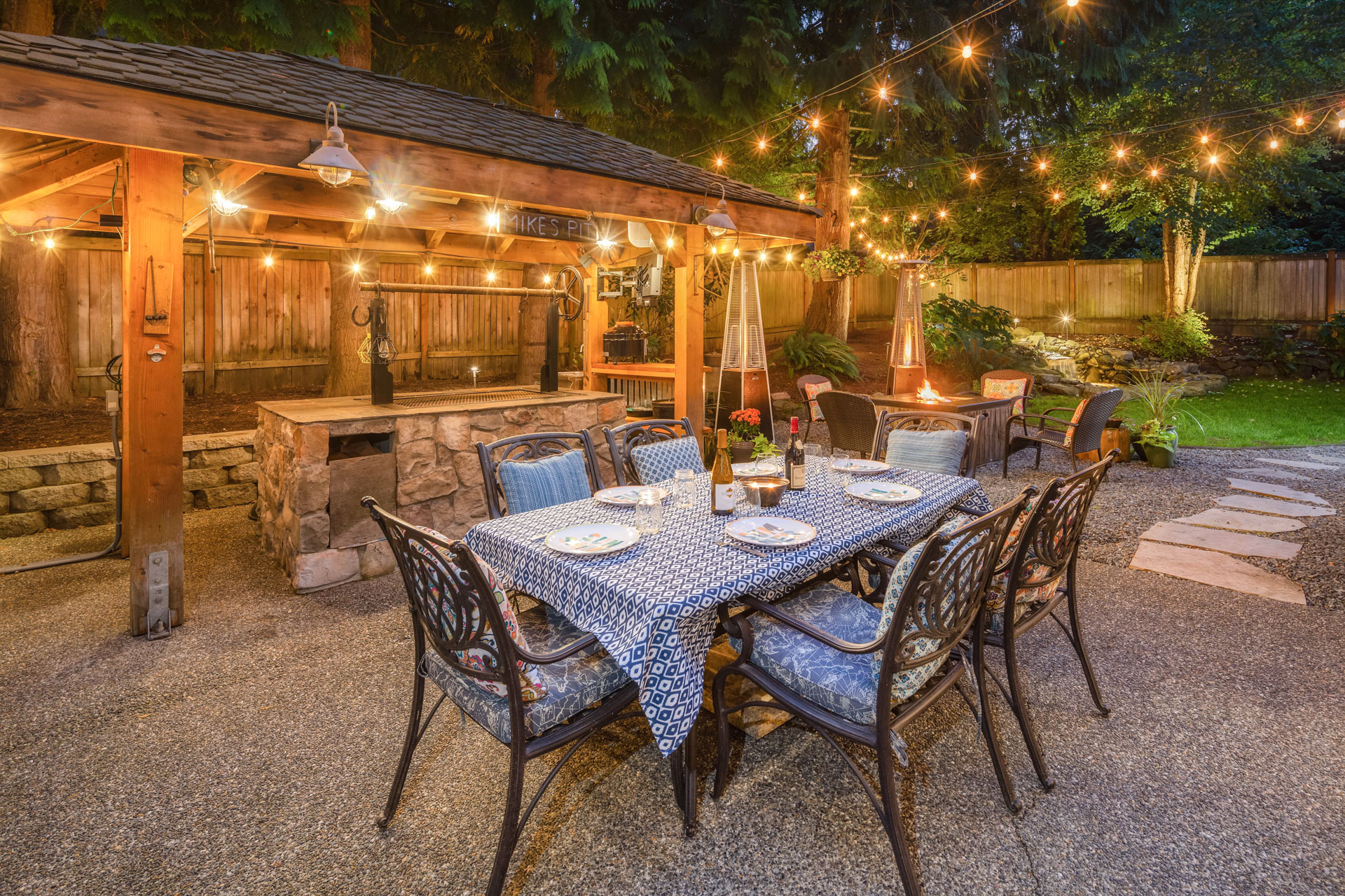 A cozy outdoor dining area is elegantly set up at dusk. A table with blue-patterned tablecloth, plates, and wine is surrounded by metal chairs. String lights hang above, and a wooden gazebo and fire pit enhance the warm, inviting ambiance.