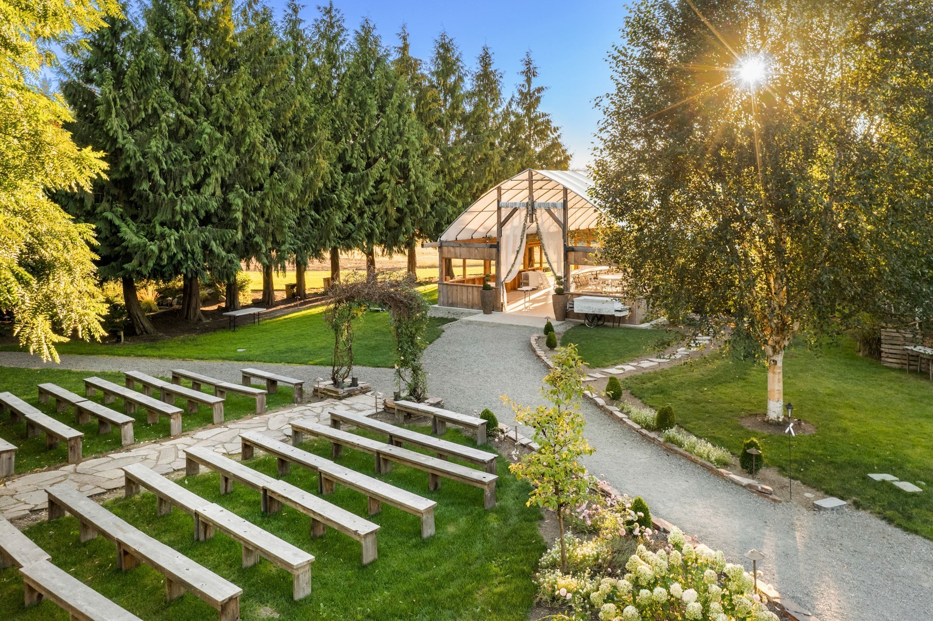 A scenic outdoor wedding venue with a gazebo in the background. The foreground features rows of wooden benches arranged on green grass, facing the gazebo. The area is surrounded by lush trees and decorated with plants and flower beds. The sun is shining brightly.