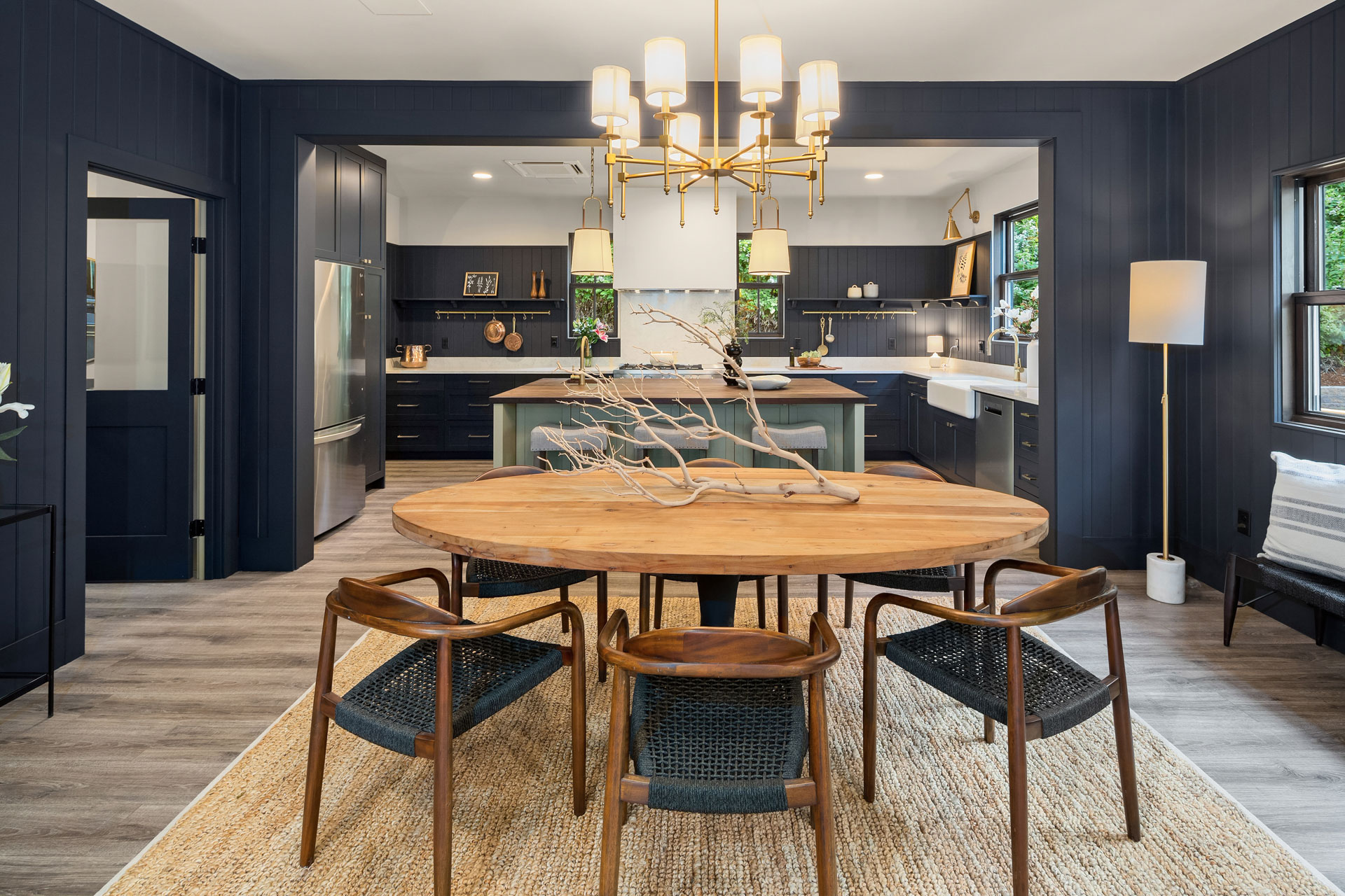 Modern dining room and kitchen with a round wooden table, four woven chairs, and a branch centerpiece. Dark blue cabinets, stainless steel appliances, and a white farmhouse sink are visible. A woven rug is under the table, and a chandelier hangs above.