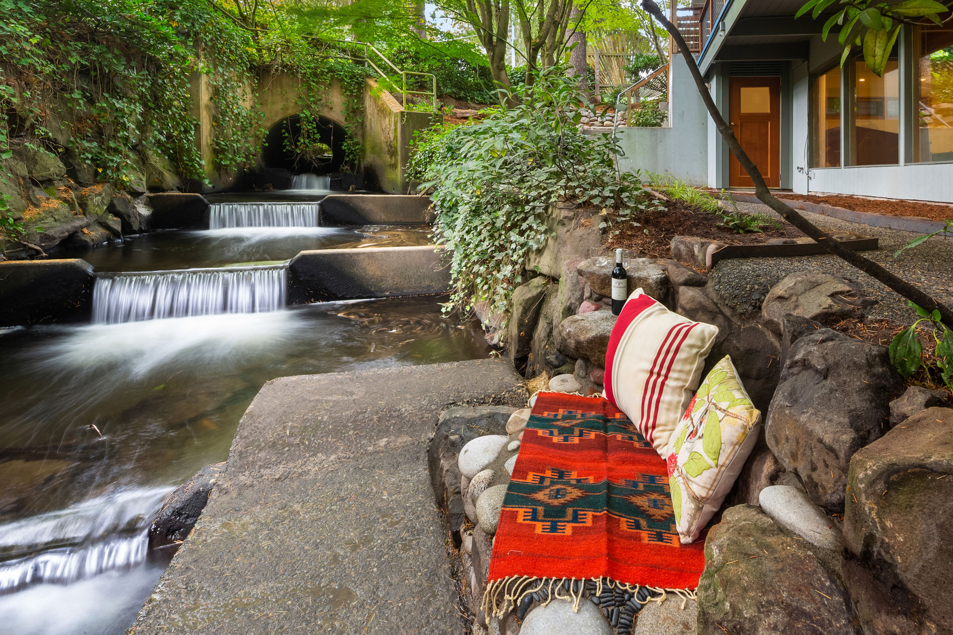 A serene outdoor seating area by a small waterfall with a woven red blanket and multicolored pillows on a stone slab. A bottle of wine and two glasses rest on a nearby rock. Lush greenery surrounds the scene, with a house visible in the background.