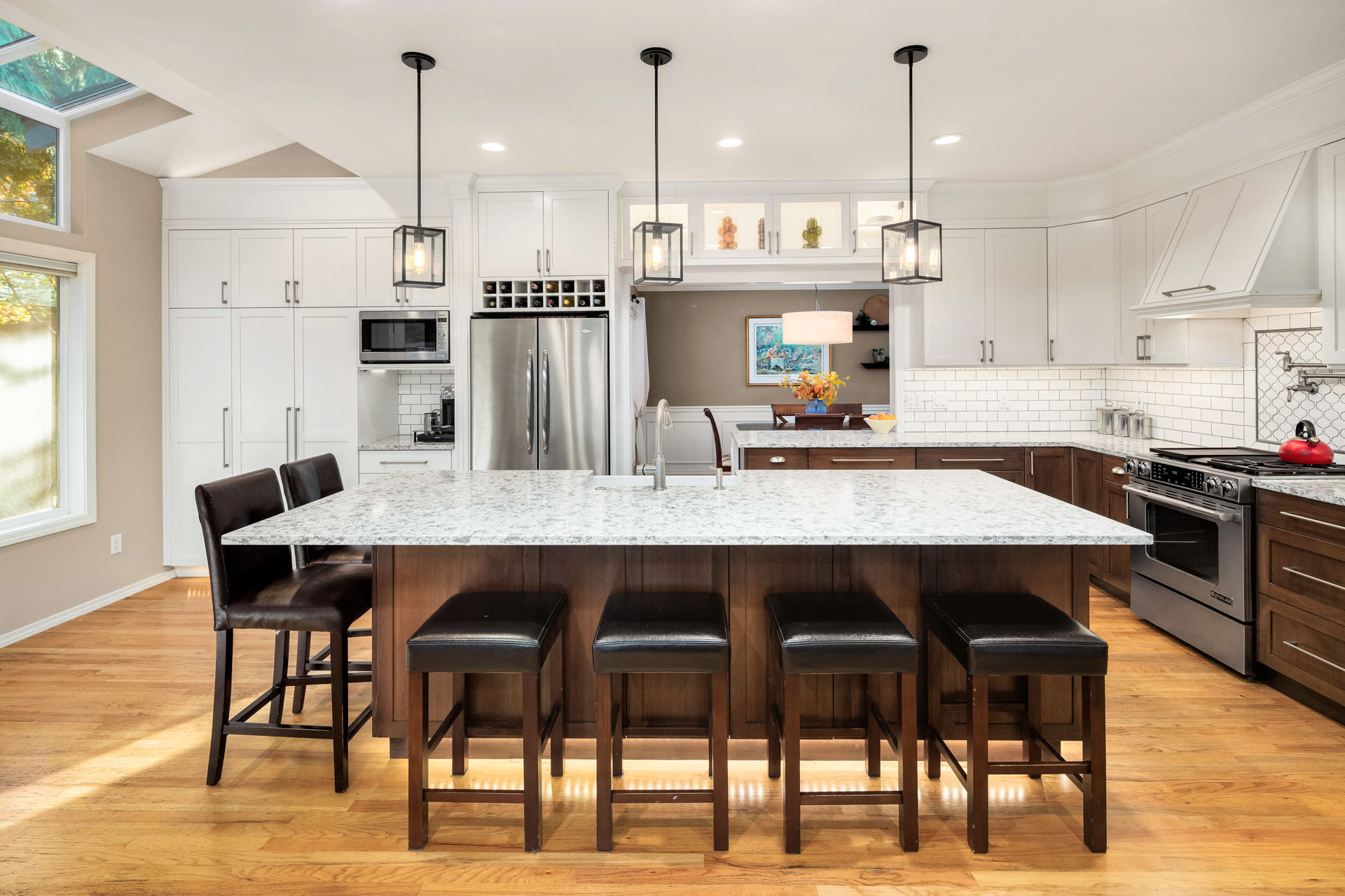 A modern kitchen features a large marble island with four wooden barstools. White cabinets with stainless steel appliances line the walls. Three pendant lights hang above the island, and a window brings natural light into the space.