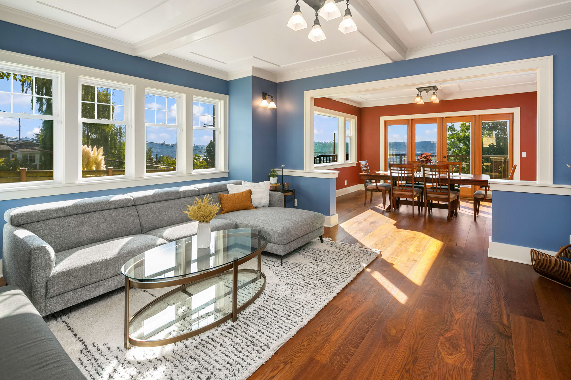 A spacious living room with a light gray sectional sofa, a glass coffee table, and a textured white rug. The walls are blue with large windows, and the dining area with red walls and wooden furniture is visible through a wide entrance. Sunlight streams in, creating a warm ambiance.