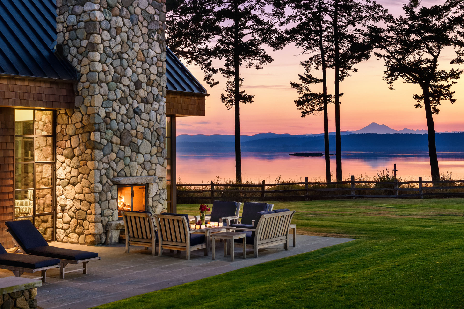 A cozy outdoor seating area with wooden chairs and a table surrounds an outdoor stone fireplace at sunset. The scene overlooks a tranquil lake with a mountainous landscape and silhouetted trees in the background, creating a serene and picturesque view.