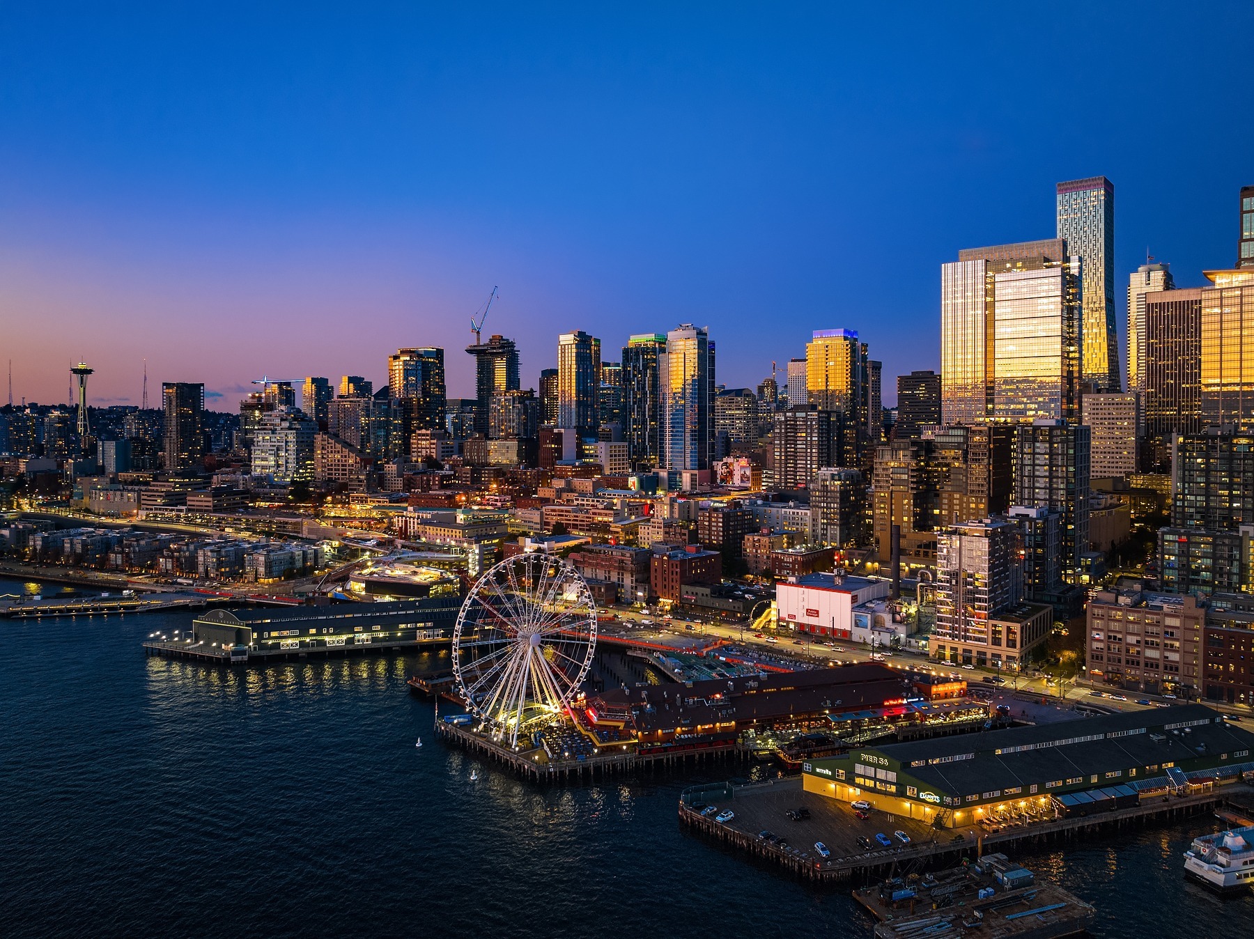 Aerial view of a cityscape at dusk featuring a waterfront with a brightly lit Ferris wheel. Tall buildings, including the iconic Space Needle on the left, are illuminated against a clear evening sky transitioning from blue to orange.