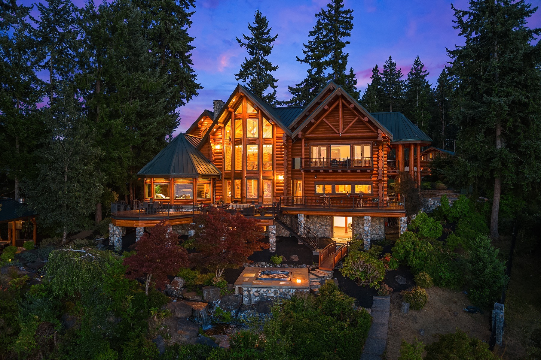 An illuminated log cabin-style home surrounded by tall trees at dusk. The multi-story house features large windows, balconies, and stone accents. A lit garden pathway leads to a small outdoor jacuzzi, all set against a vibrant purple and pink sunset sky.