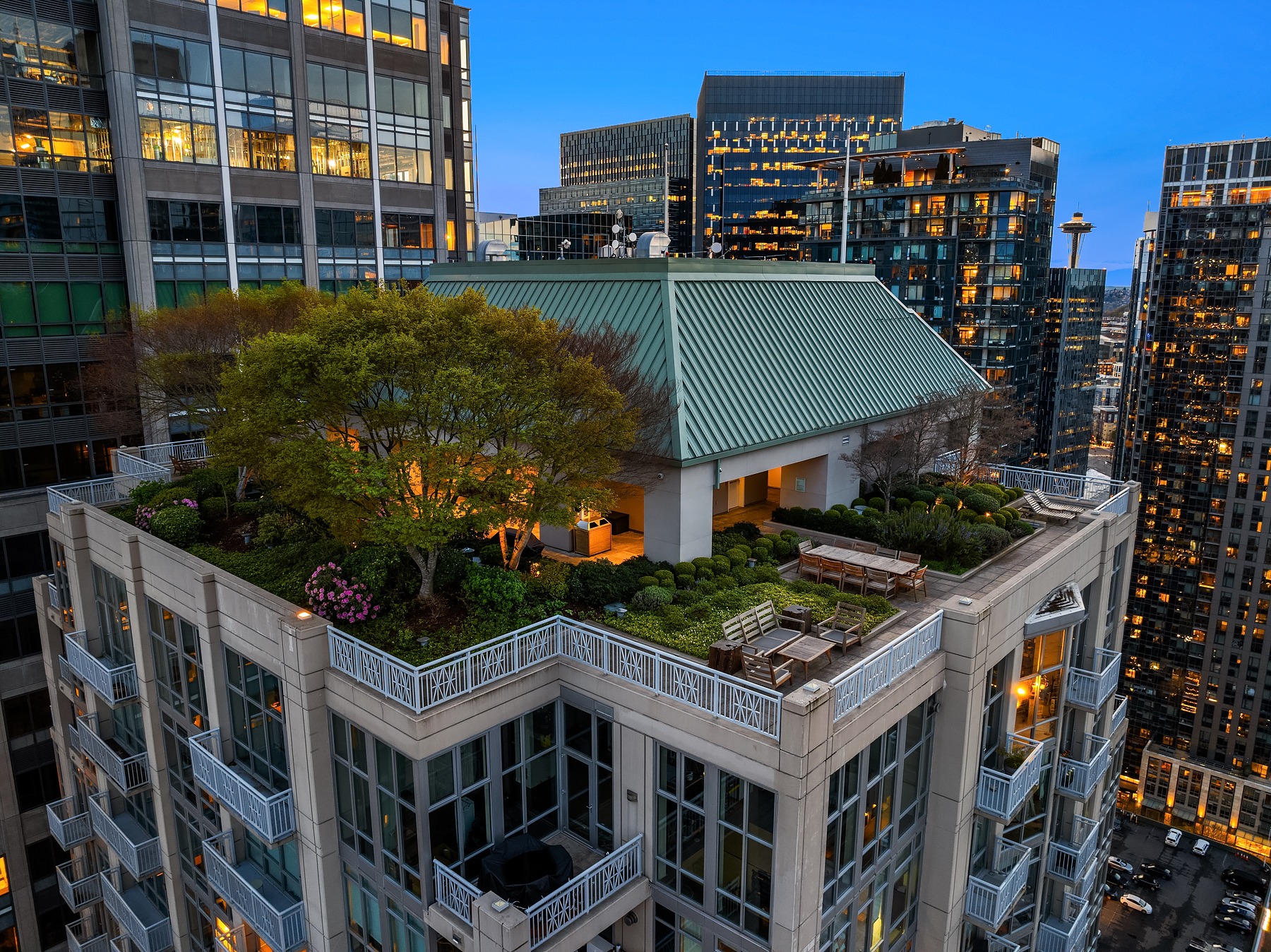 A rooftop garden with trees, shrubs, and a seating area sits atop a modern high-rise building at dusk. Surrounding buildings with large windows reflect the evening sky. City skyline and illuminated tower can be seen in the background.