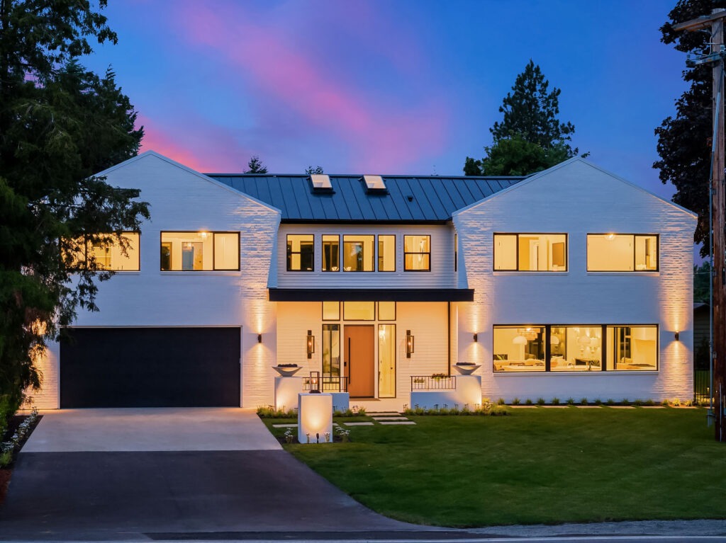 Modern two-story house with large windows and a sleek design, illuminated at dusk. The exterior is white with a dark roof and features a spacious driveway leading to a double garage. The well-maintained lawn and ambient lighting enhance the contemporary aesthetic.