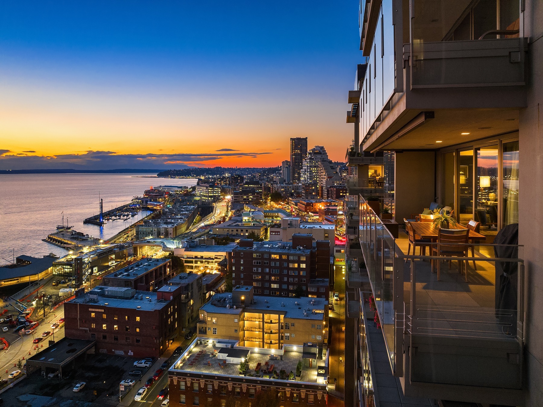 Aerial view of a city at sunset, with buildings and streets lit up. A balcony on the right features a table set for two with chairs, overlooking the cityscape and waterfront. The sky transitions from orange near the horizon to deep blue as night falls.