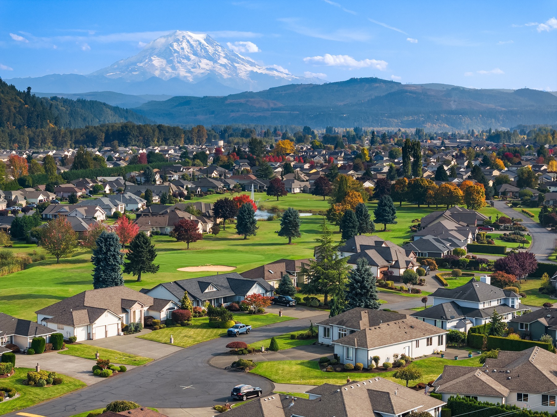 Aerial view of a suburban neighborhood with neatly arranged houses and green lawns. The scene is set against a backdrop of mountains with snow-capped peaks, under a clear blue sky. Trees with autumn foliage add vibrant color to the landscape.