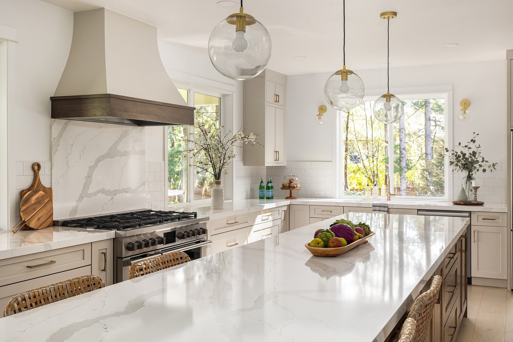 Modern kitchen with white marble countertops and light beige cabinets. A large center island features decorative glass light fixtures above. Stainless steel appliances and a wooden cutting board are visible. There are vases with flowers and a bowl of fruits.