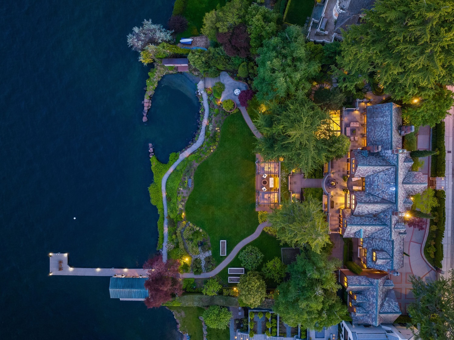 Aerial view of a large lakeside property featuring an illuminated house with an expansive garden, winding pathways, a small dock, and lush greenery. The property is bordered by calm blue water and is surrounded by various trees and plants.