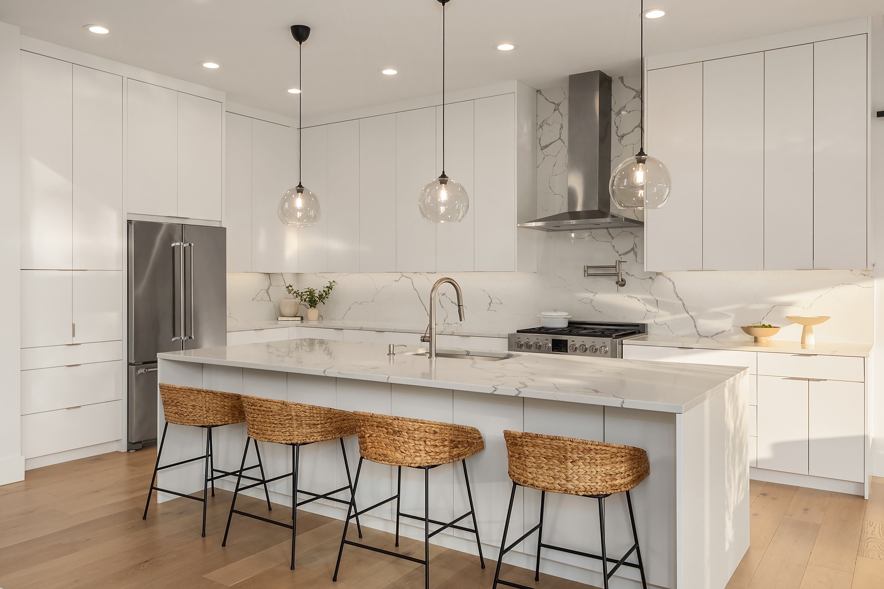 Modern kitchen with white cabinets, marble countertops, and a center island. Three pendant lights hang above the island, which has four woven stools. The kitchen features stainless steel appliances, wooden flooring, and a minimalist decor with subtle greenery.