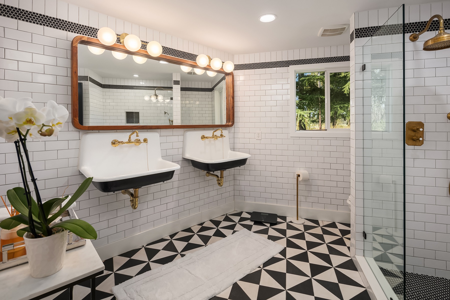 A modern bathroom with white subway tile walls, black and white geometric floor tiles, and a large mirror above two sinks. There is a glass shower enclosure, potted orchids on a table, a small window, and bright vanity lights.