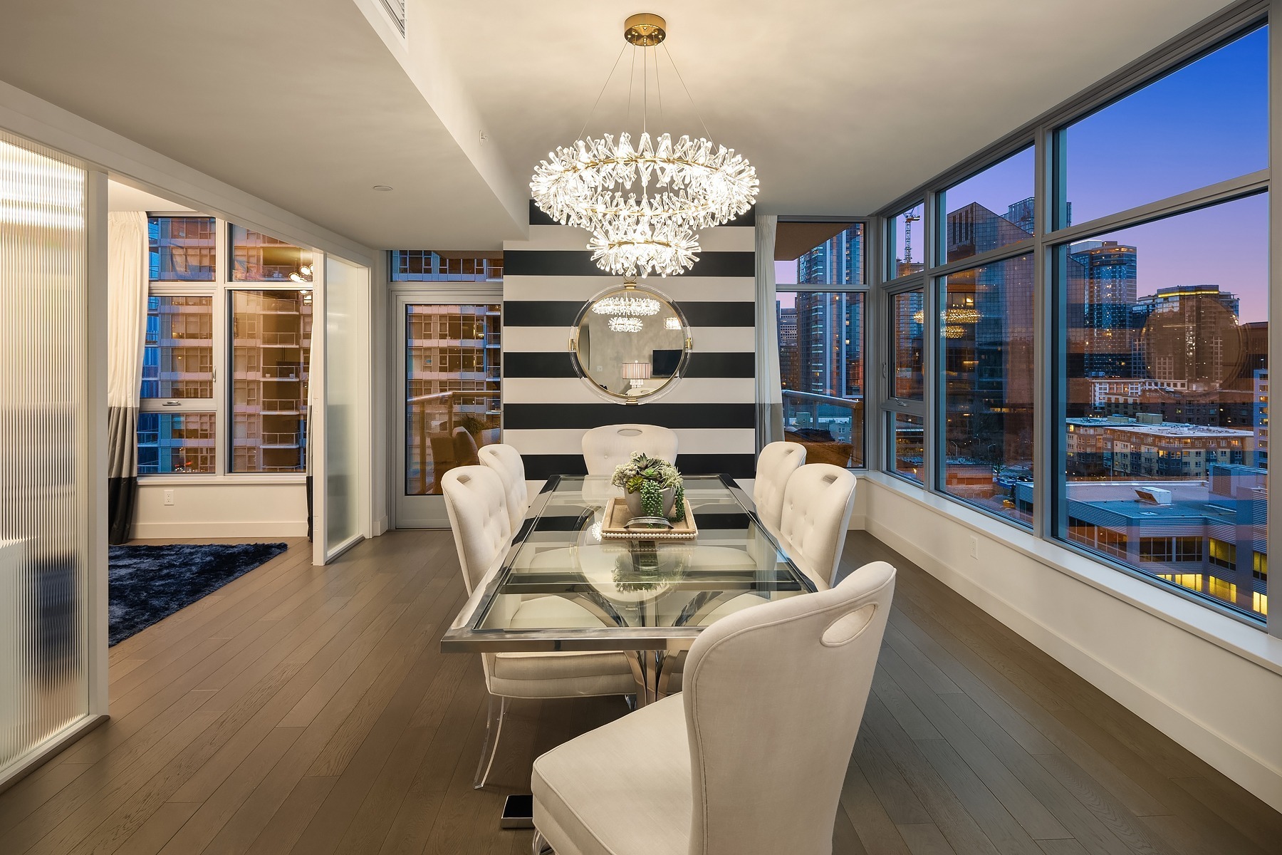 Modern dining room with a large glass table surrounded by white chairs. A striking chandelier hangs above the table, and large windows offer a view of a city skyline at dusk. The walls feature bold, black and white stripes, and a round mirror is mounted on one wall.