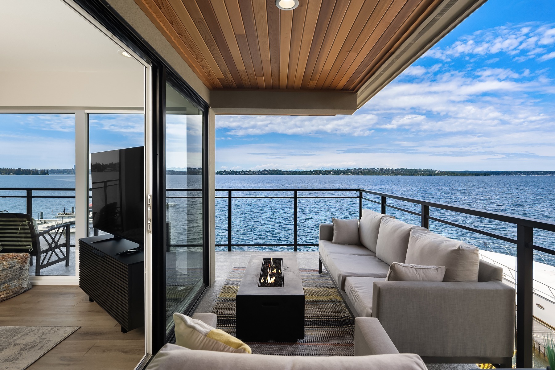 A modern outdoor balcony with a wooden ceiling features a cozy seating area with a grey sectional sofa and a fire pit table. It overlooks a large body of water. Sliding glass doors connect the balcony to the indoor living area with a flat-screen TV.